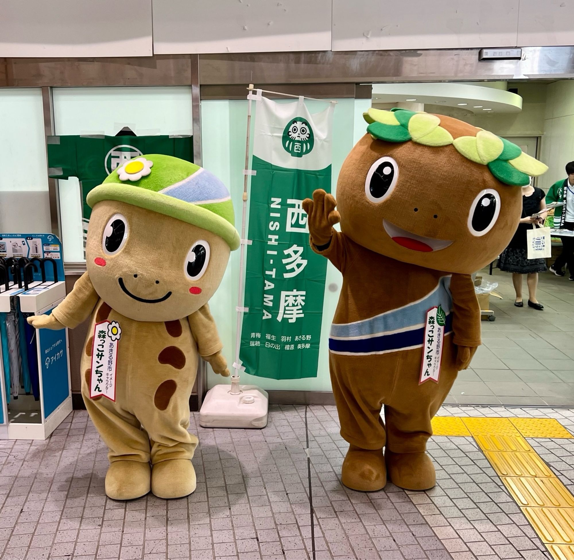 A small beige salamander mascot with a green hat stands beside a large brown salamander in a train station.