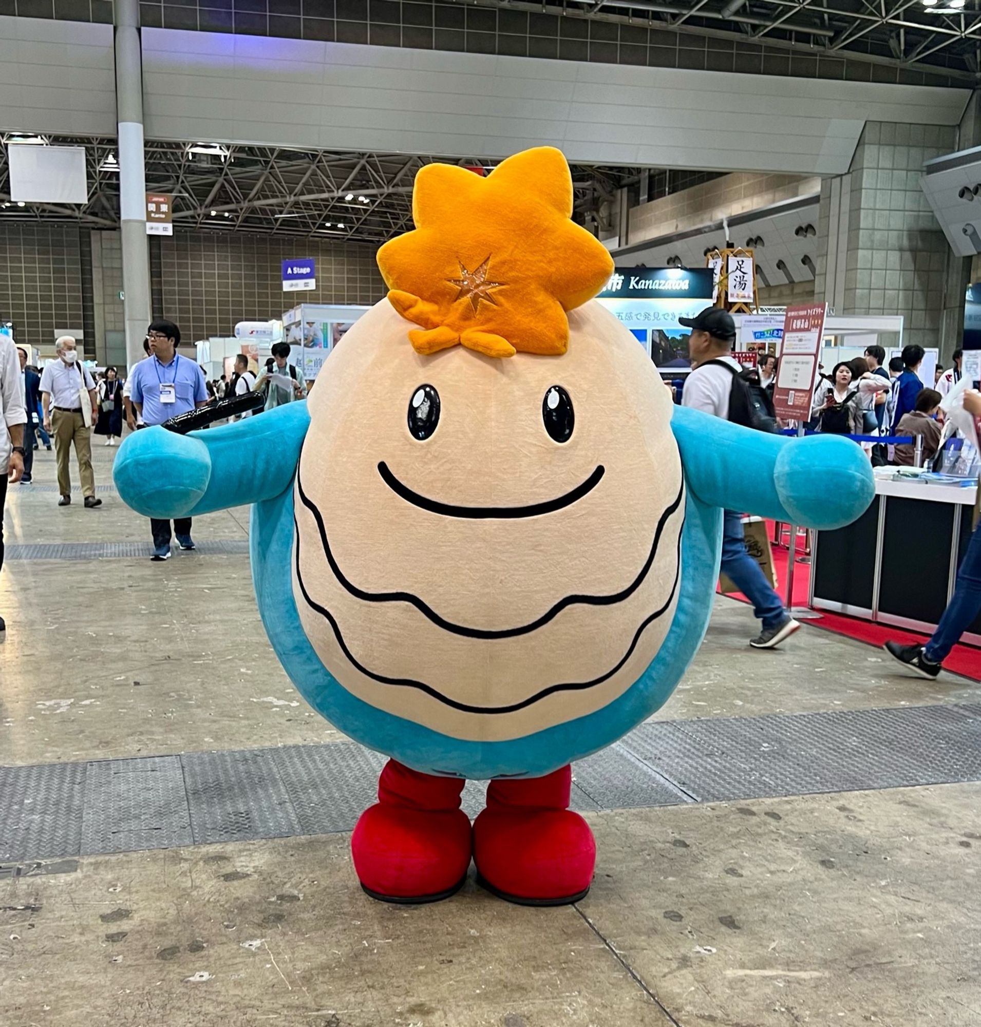 An oyster mascot with a maple leaf on its head stands, arms outstretched, in a convention center.