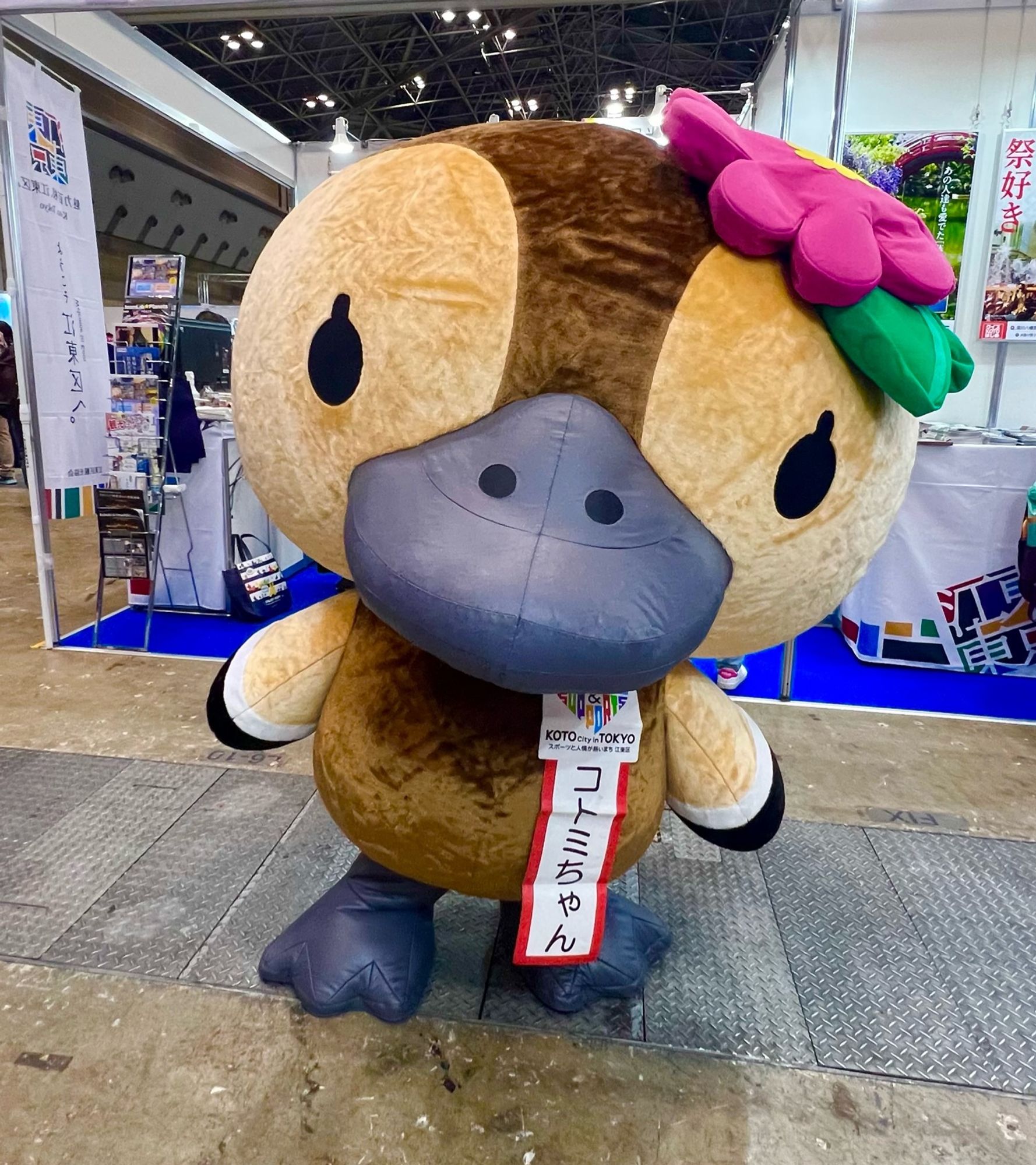 A brown duck mascot with a camellia in its hair poses for the camera.