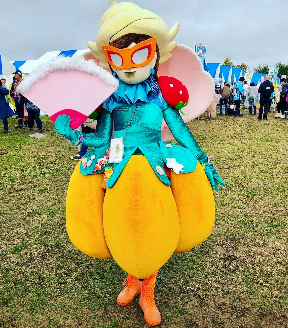 A fairy whose bottom half is a loquat holds a pink fan and wears big orange glasses and stands in a field.