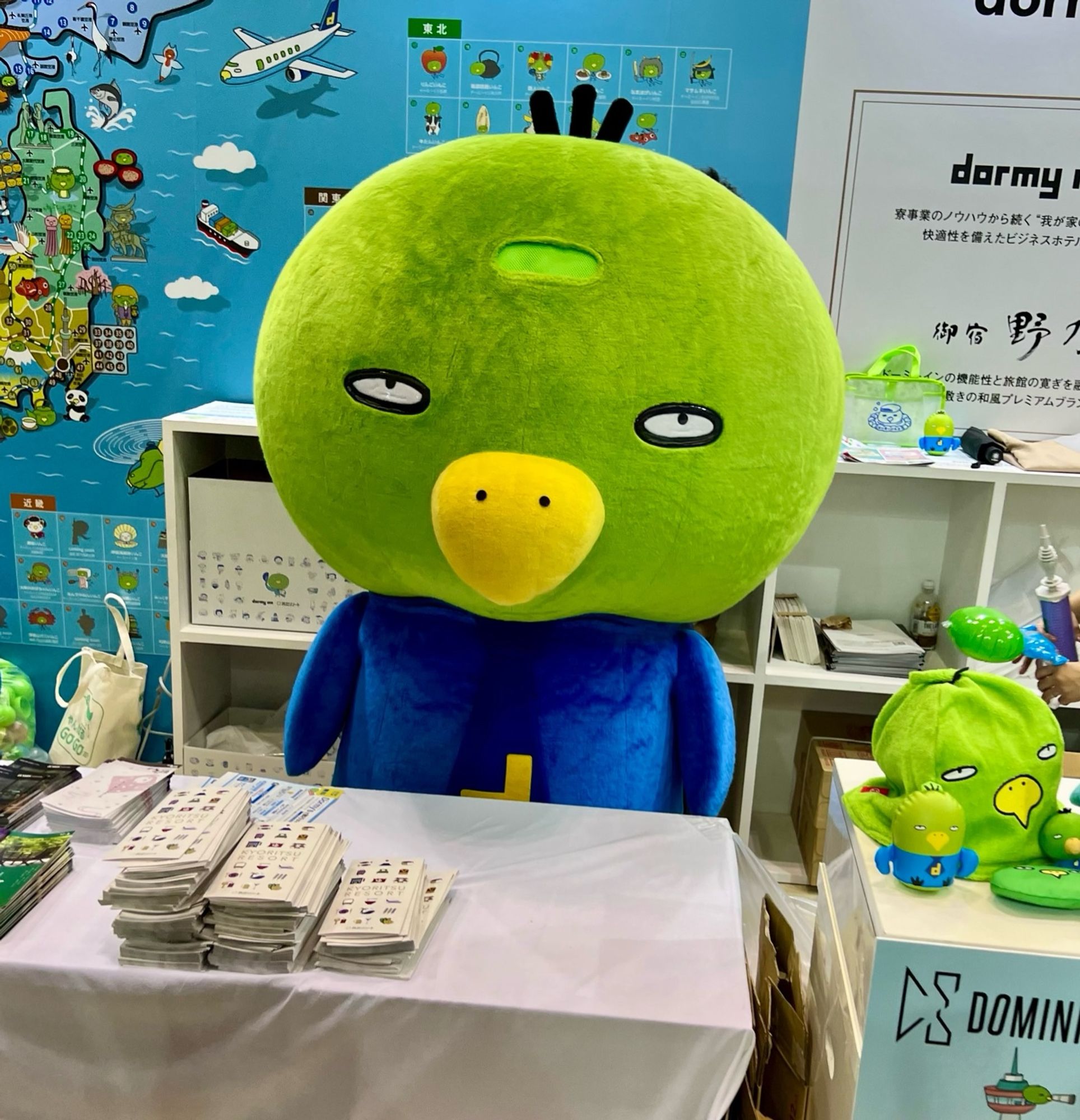 A green parakeet mascot in a blue shirt sits at a table in a convention centre.