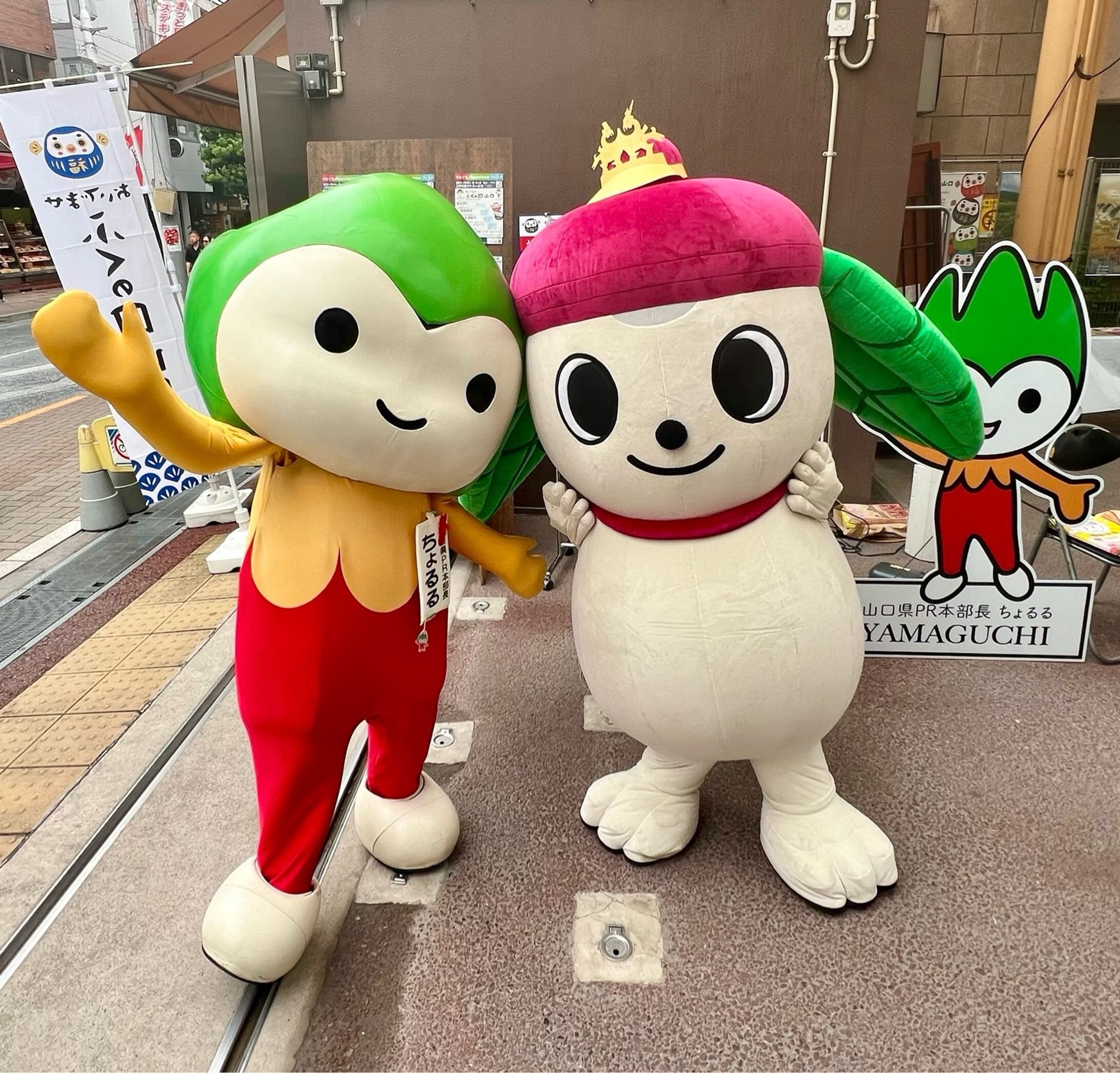 A mascot with green hair, red pants, and an orange top poses with a dog mascot with a tea bowl hat and green tea leaves for ears.