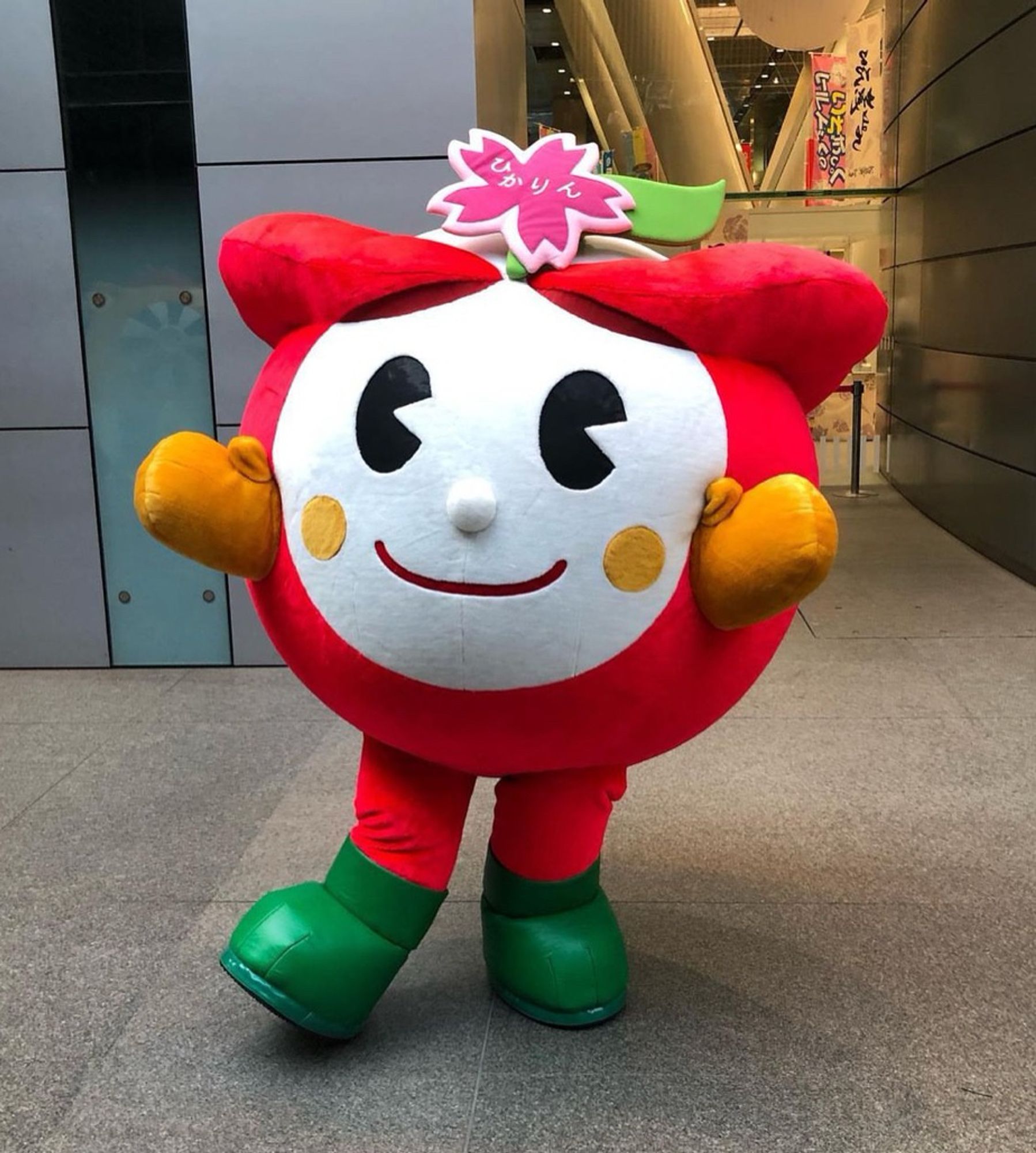 A spherical red mascot with a white face, green boots, and a pink flower in its hair poses for the camera.