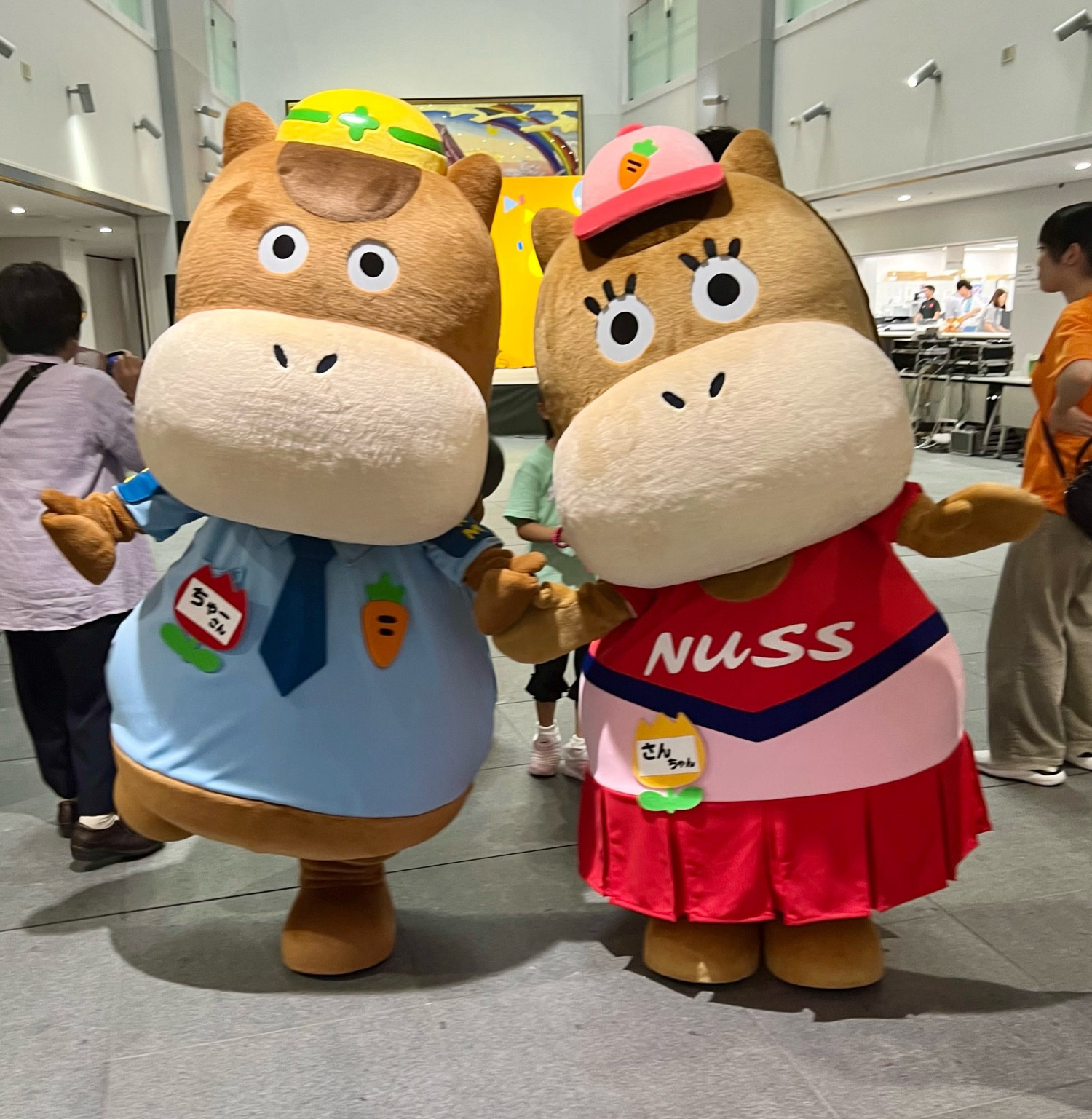 Two horse mascots wearing caps, one in a blue shirt and tie and the other in a red and pink dress, pose in a university lobby.