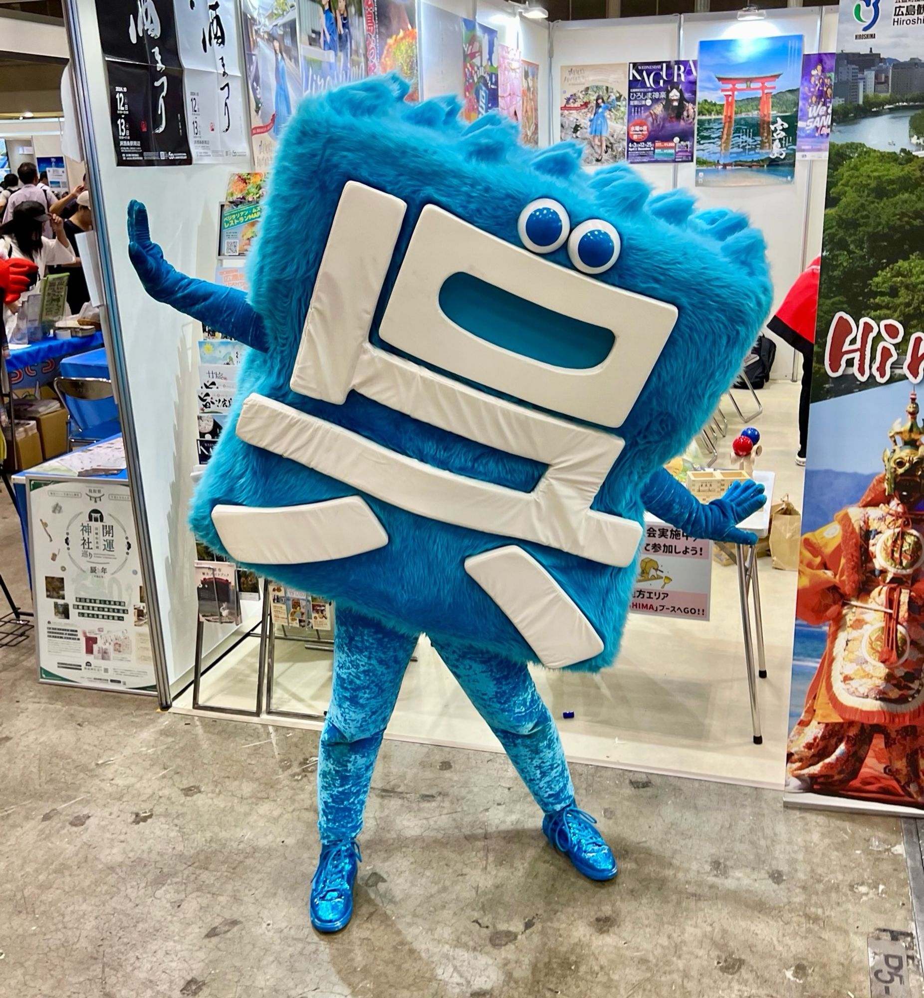 A square blue mascot, with the Japanese kanji “Kure” on its chest, poses daintily in front of a tourism promotional display.