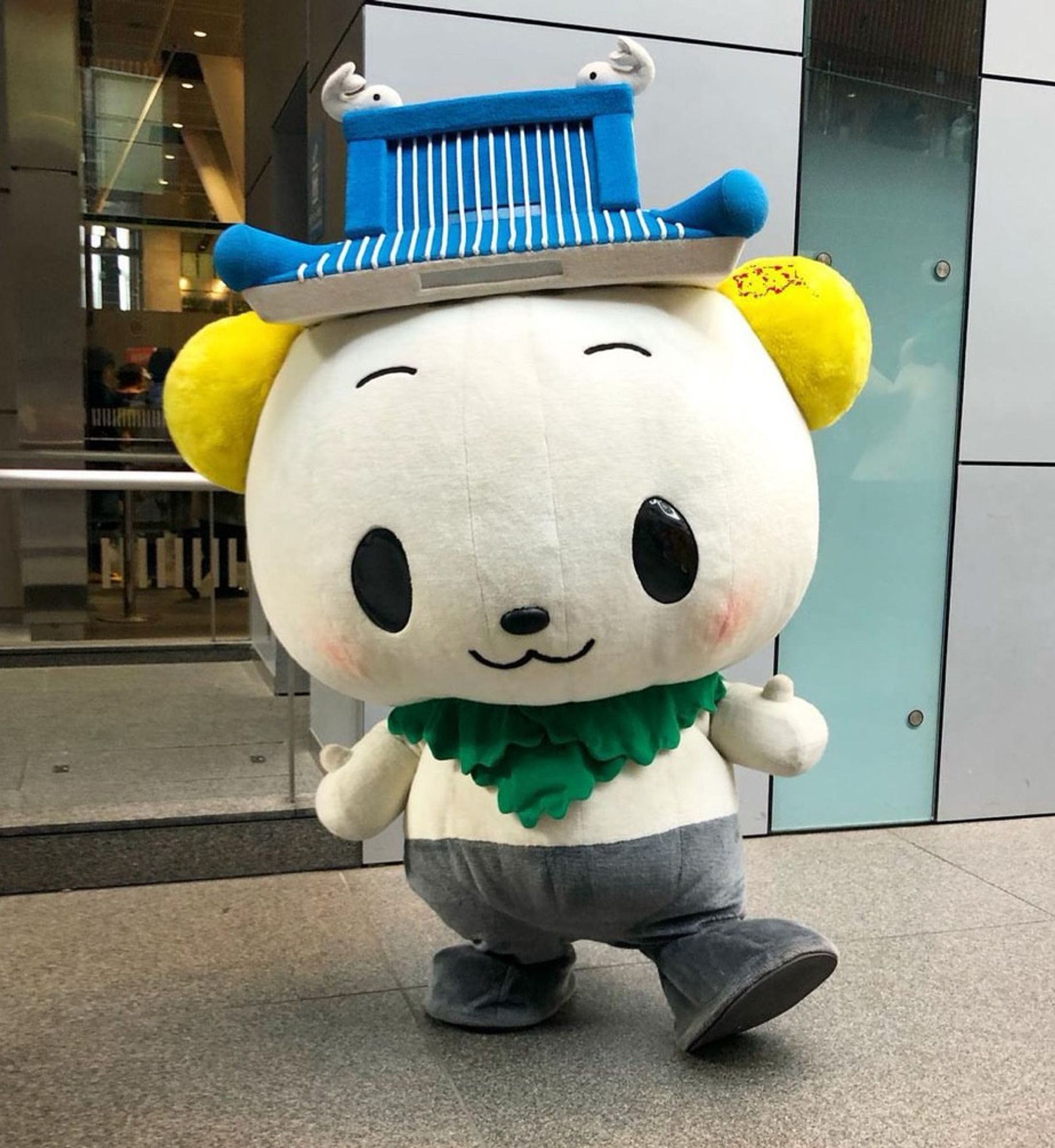 A mascot stands in a convention center. He has a castle hat with a couple of whitebait on top, chrysanthemum earrings, and a lettuce scarf.