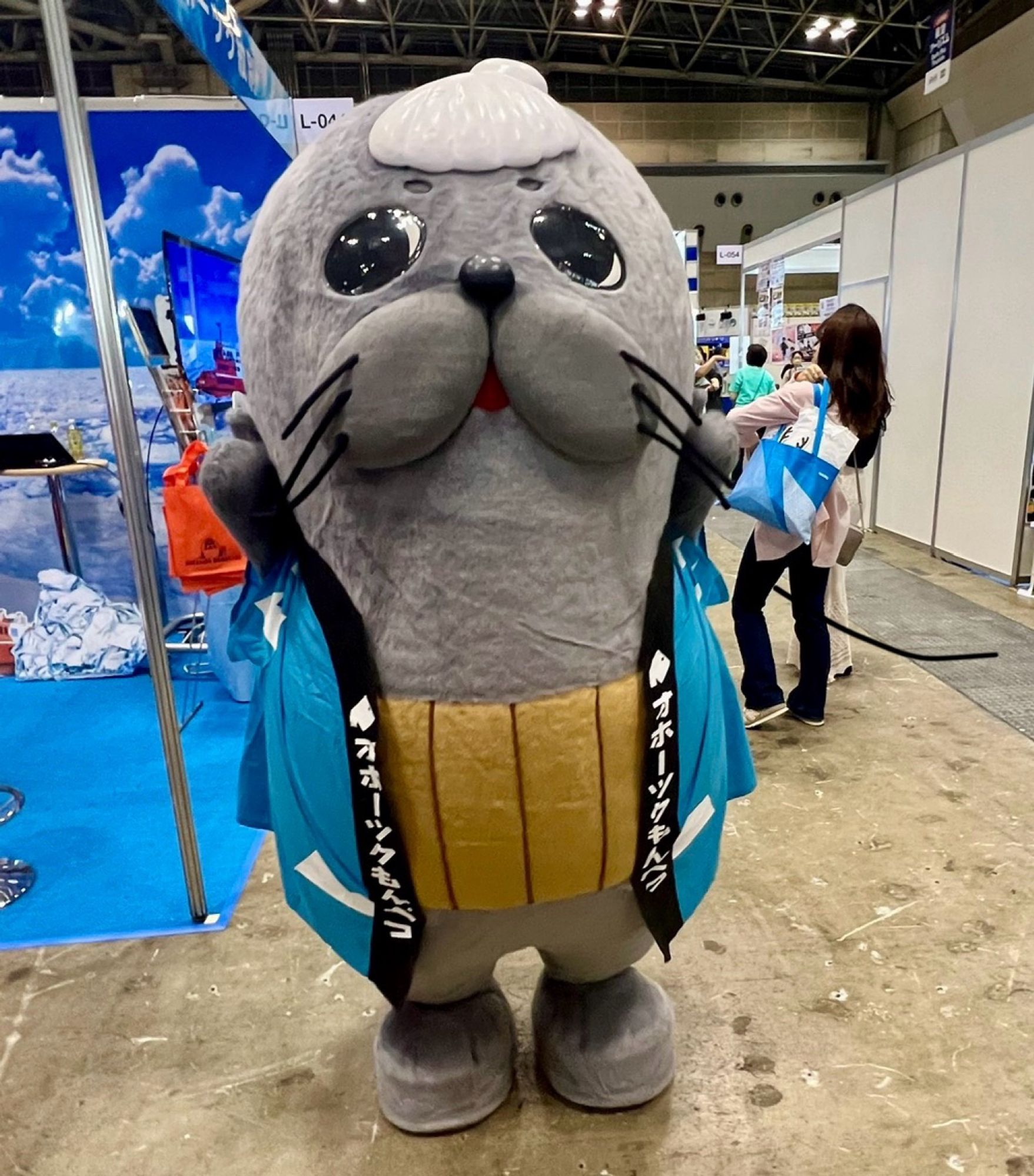 A seal with a scallop shell balanced on its forehead wears a blue jacket and stands in a convention centre.