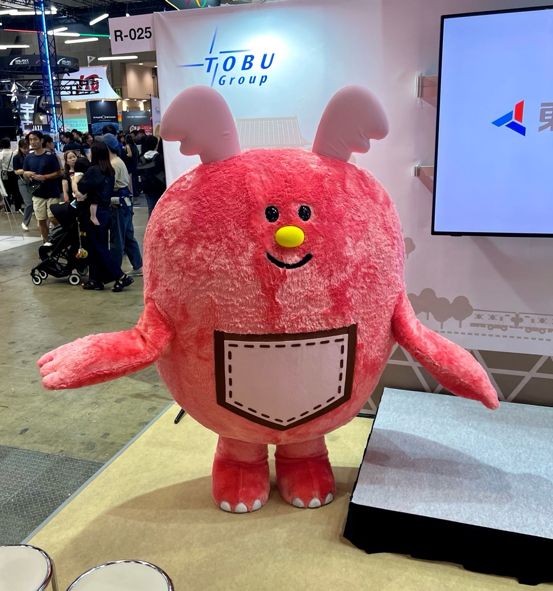 A pink monster with wing-like ears and a large pocket on his belly stands beside a stage.