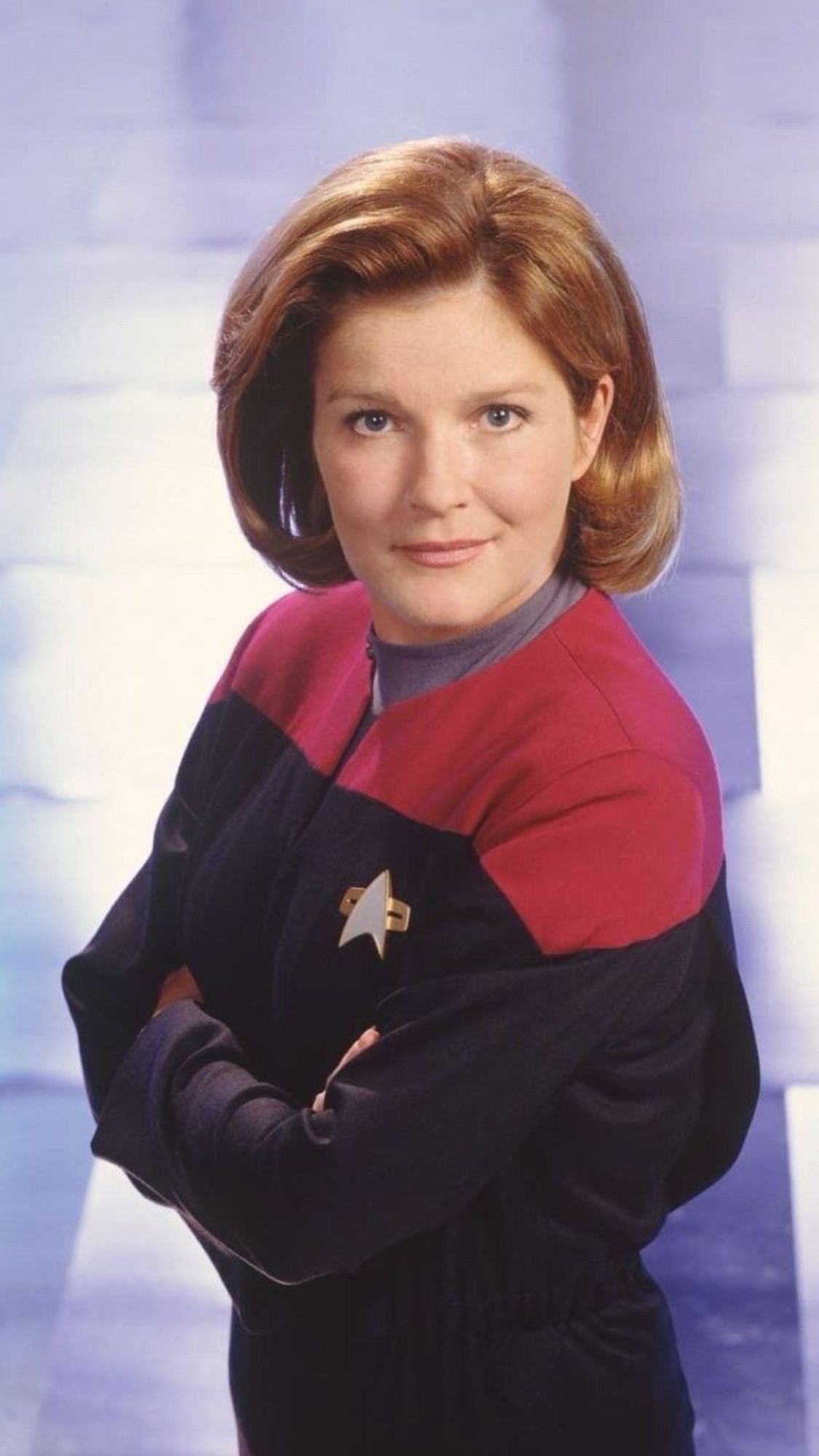 Woman in red Star Trek uniform, captain Janeway. Redish hair in a to the shoulders bob and her arms crossed over her chest, a wry smile on her face.