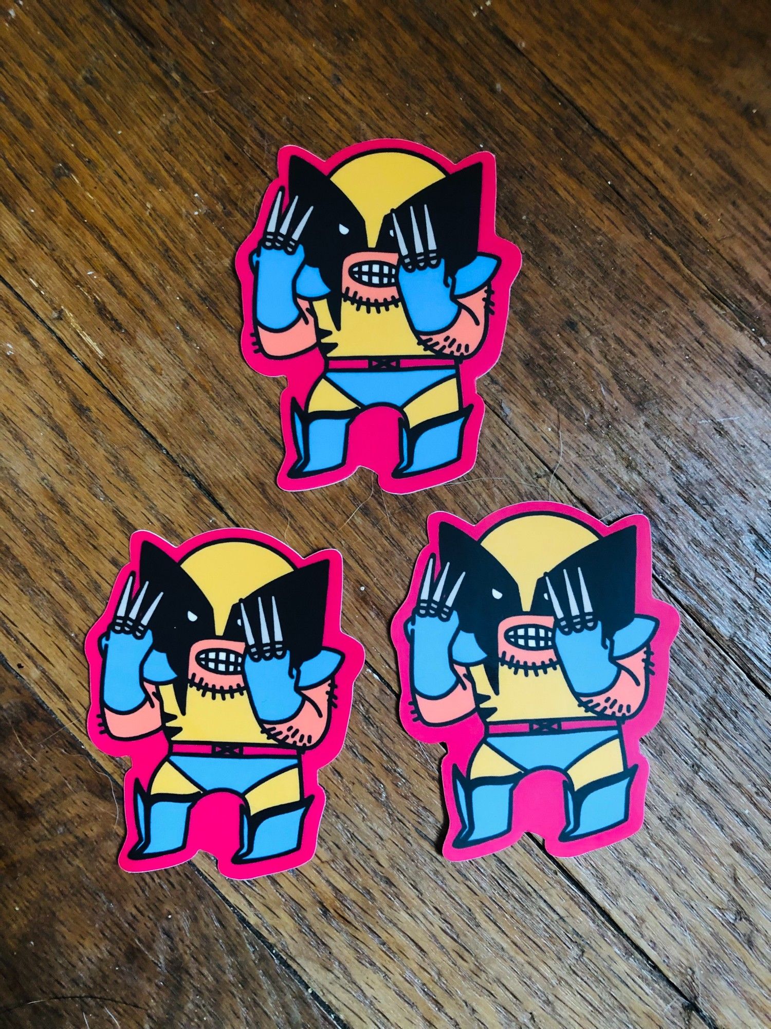 it's 3 stickers of a little tiny wolverine, holding his claws in front of his little face. they are on a wooden background.