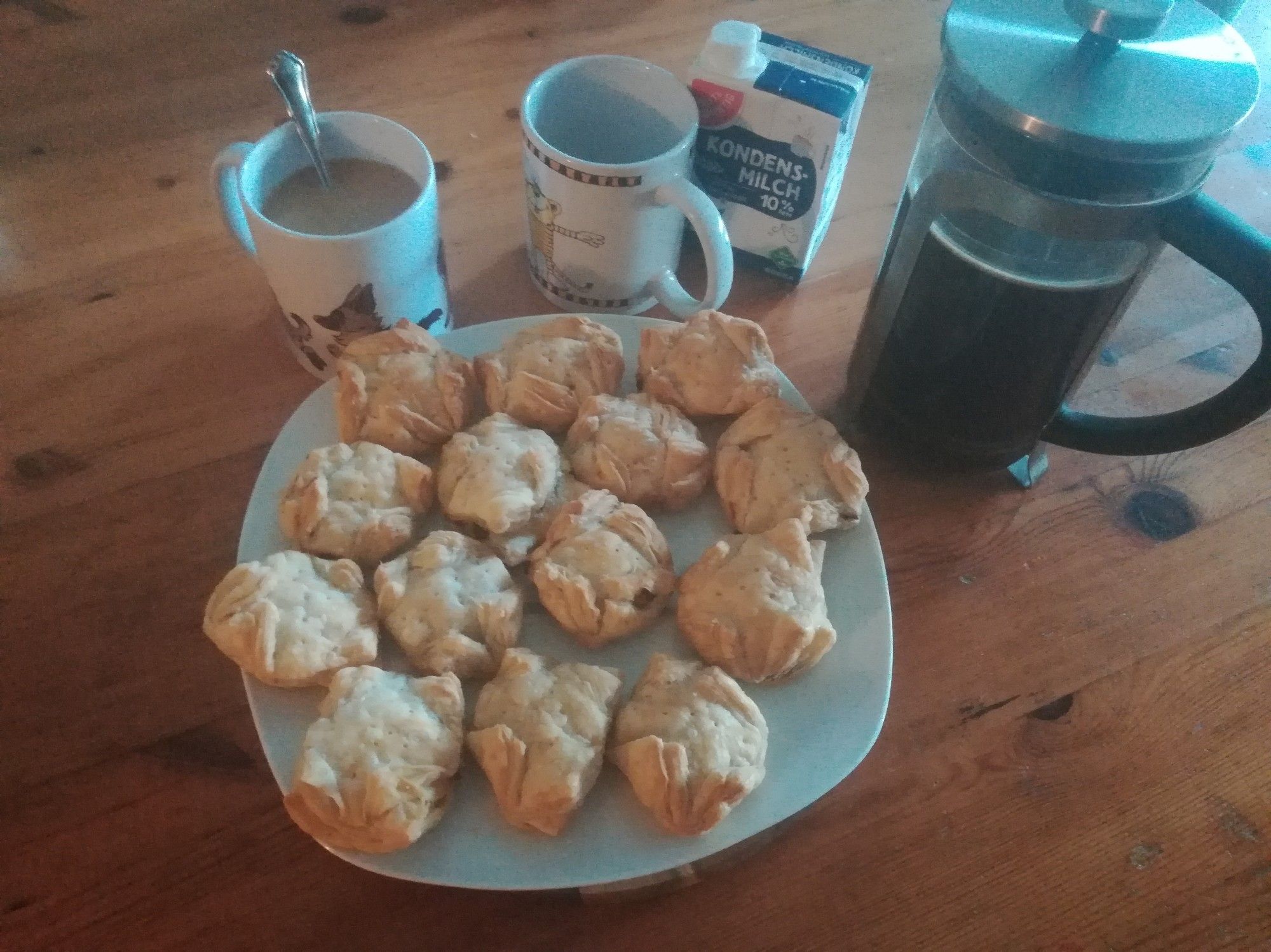 Homemade puff pastry pockets filled with onions, cream cheese, cheddar, gouda and garlic. Next to it is a coffee in a cup with a cute hyena pattern and an empty cup with Janosch. Plus a French press with fresh coffee and a carton of condensed milk.