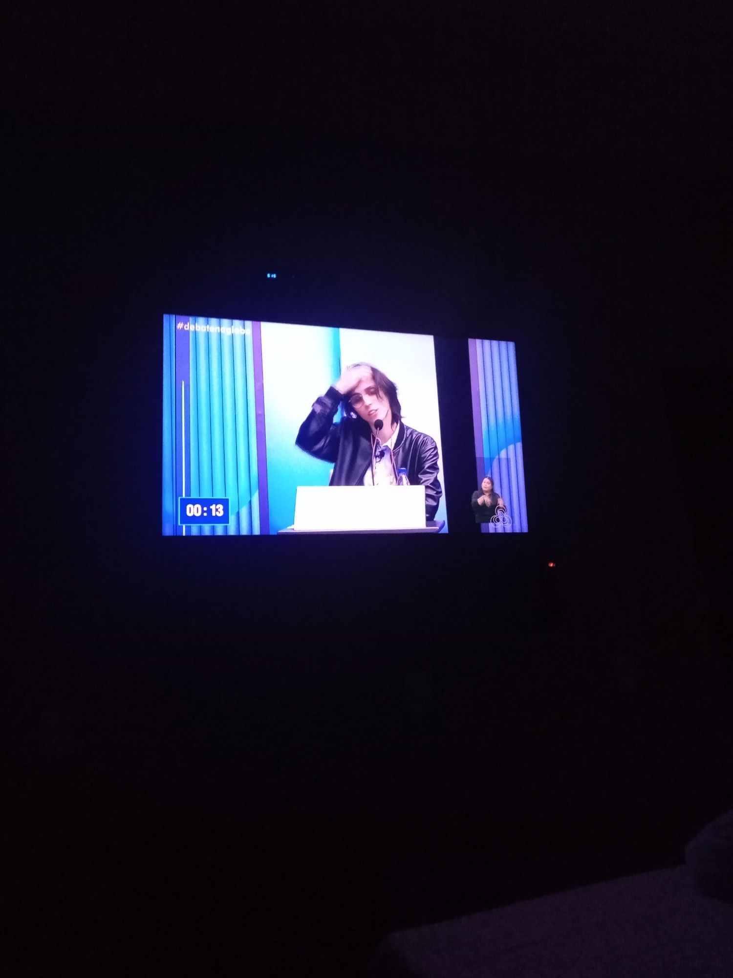 Foto de televisão onde Amom Mandel no debate da Globo está bagunçado os cabelos com um timer na tela. Ele é um homem branco, magro, com óculos e cabelos pretos e lisos na altura dos ombros com uma franja. Ele usa óculos redondos, jaqueta preta e blusa branca.