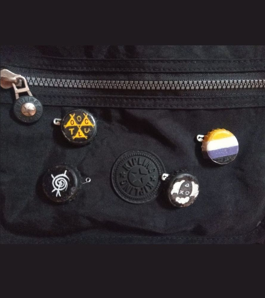 Bolsa preta no fundo com 4 bottoms de tampinhas de garrafa de vidro pintadas como: símbolo Oculto de Ordem Paranormal; bandeira não binária; símbolo do elemento Morte; símbolo da Ahlevo.