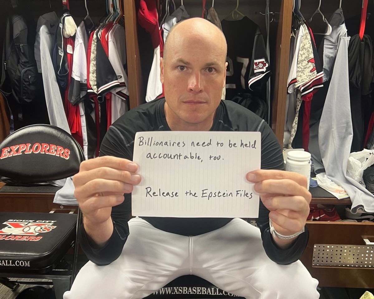IA State Representative (D., District 1)  &  US Senate Candidate JD Scholten sits on a bench in a baseball locker room wearing his pitcher’s uniform while holding a sign “release the Epstein Files.” 