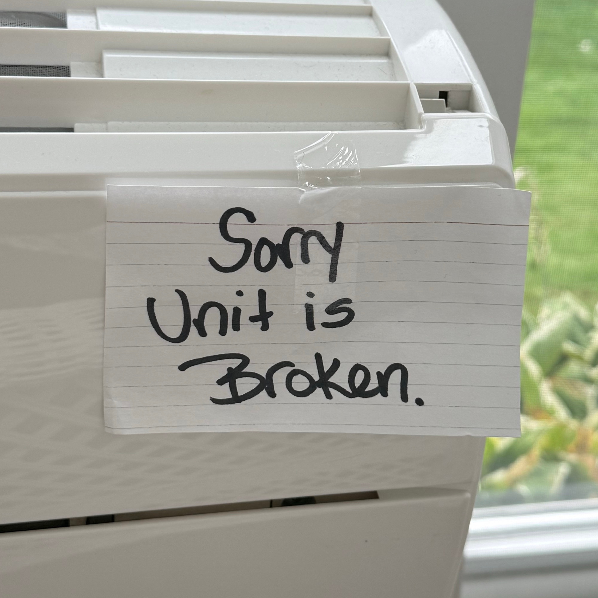 Photo of an A/C unit with an index card taped on the front which reads: Sorry Unit is Broken. 