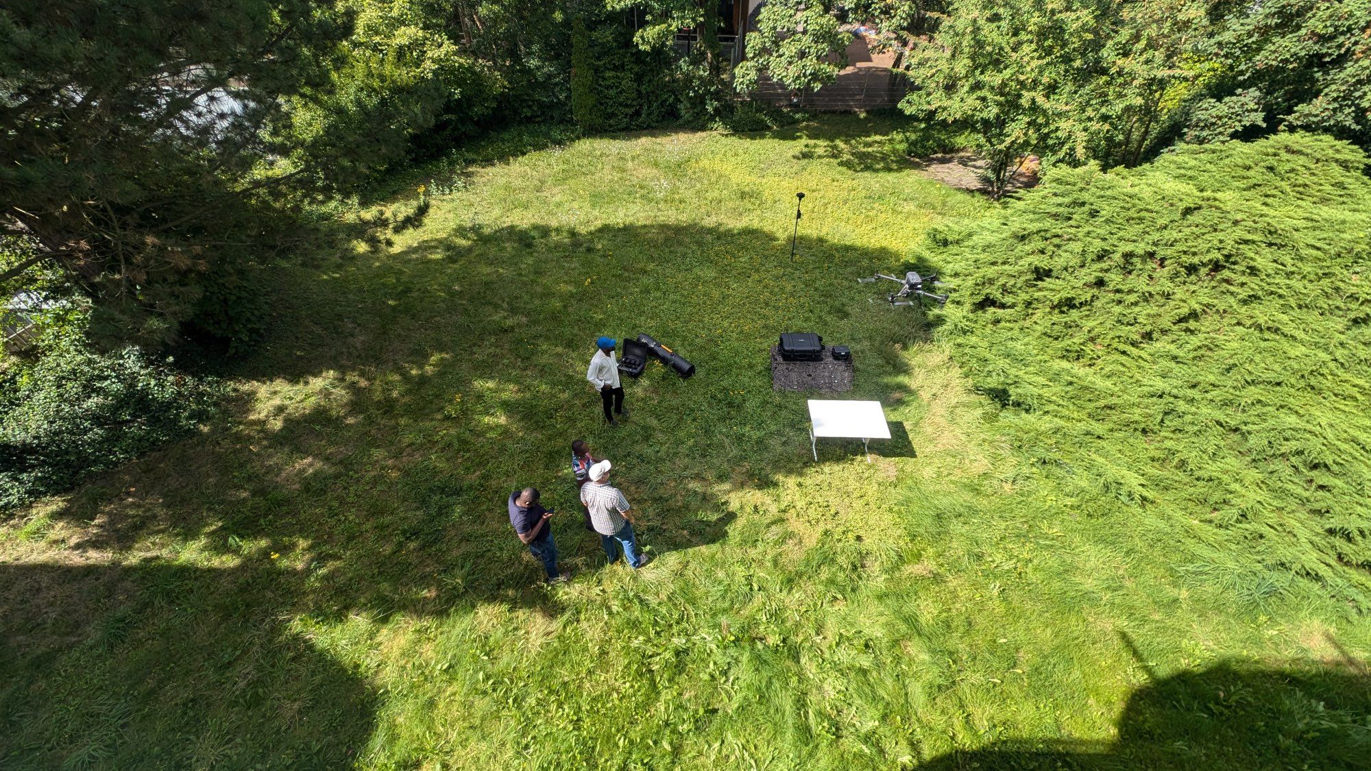 Aerial photo showing the perspective of a drone looking down at three people in a green landscape, handling another drone.