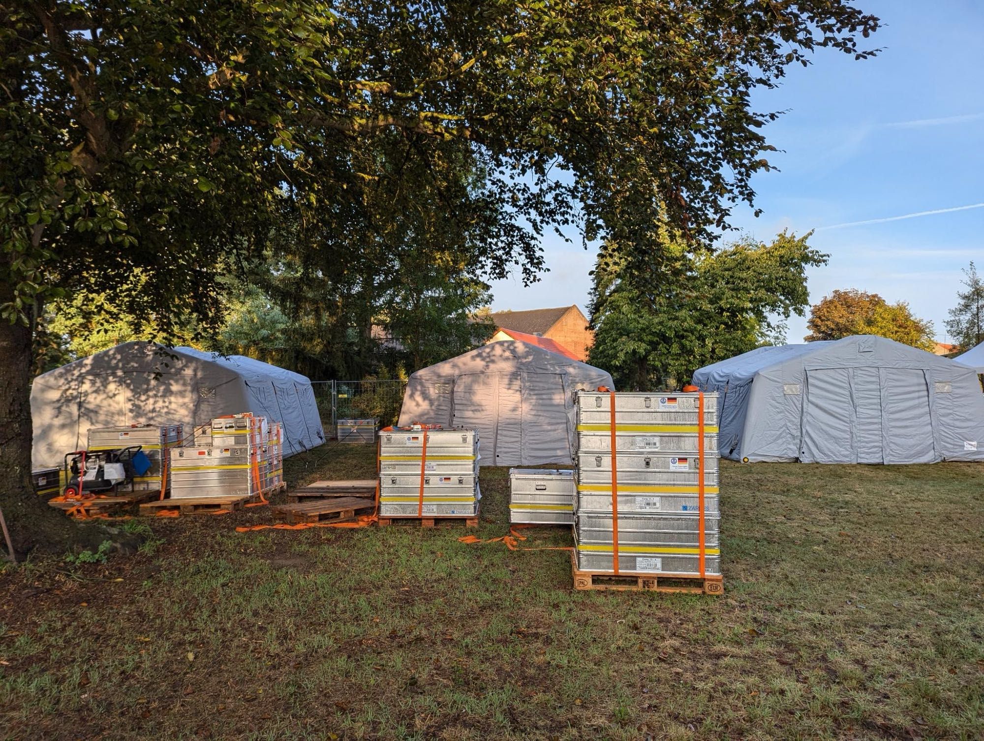 Foto dreier großer Einsatzzelte unter Bäumen; davor mehrere Aluminiumkisten, z.T. mehrere übereinandergestapelt, auf Holzpaletten.