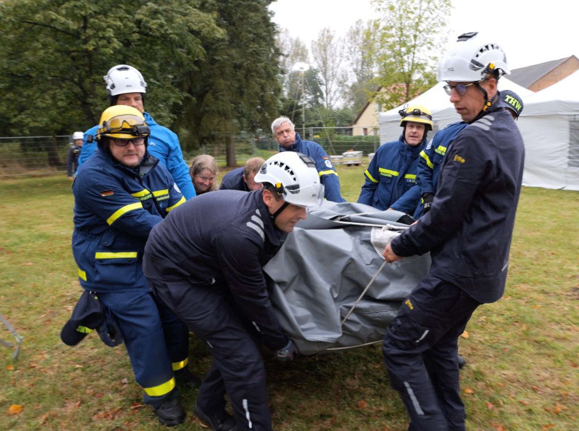 Foto einer Gruppe Personen in THW-Einsatzkleidung und Schutzhelmen beim Transport eines schweren in eine Plane gehüllten Gegenstands. Im Hintergrund sind Behelfszelte und Bäume zu erkennen.