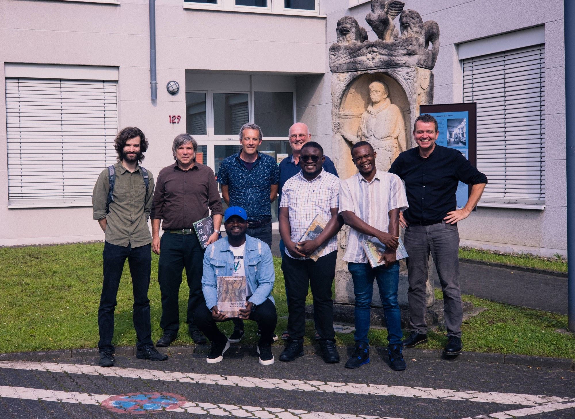 Group photo of eight men posing next to a historic monument standing in front of a modern building.