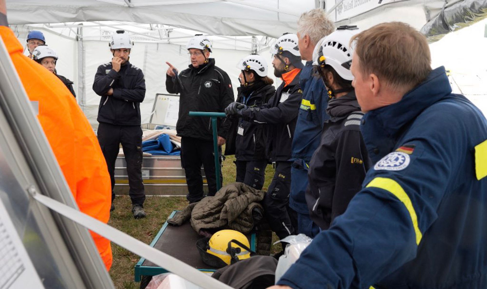 Foto einer Reihe Personen, z.T. in blauer THW-Einsatzkleidung und mit weißen Helmen ausgestattet, stehend, in einer Besprechungssituation.