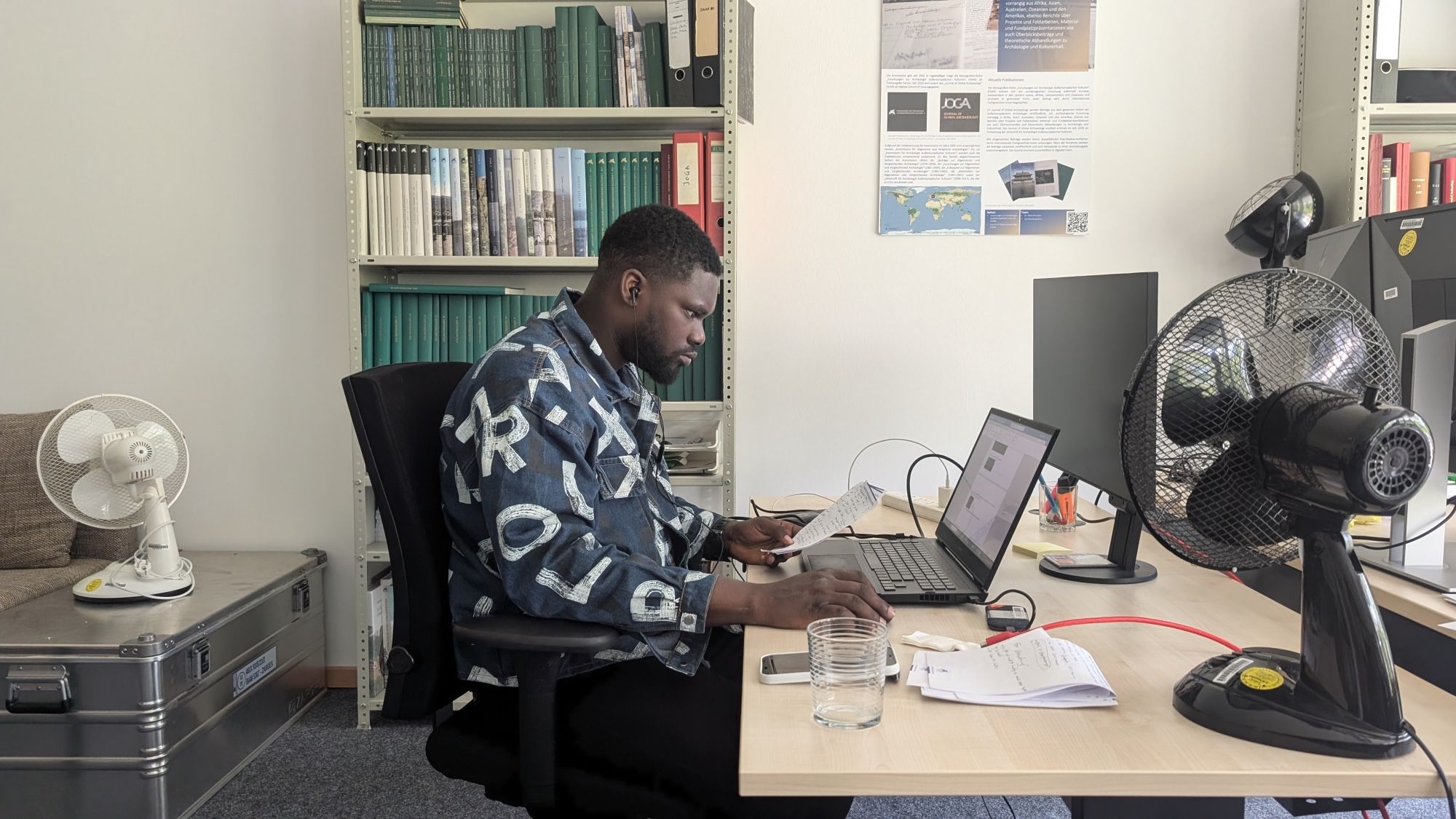 Photo showing a man sitting at the desk, looking at his notebook computer. Bookshelves and a metal transport box with a fan can be seen in the background. Another fan is standing to the right on the desk.