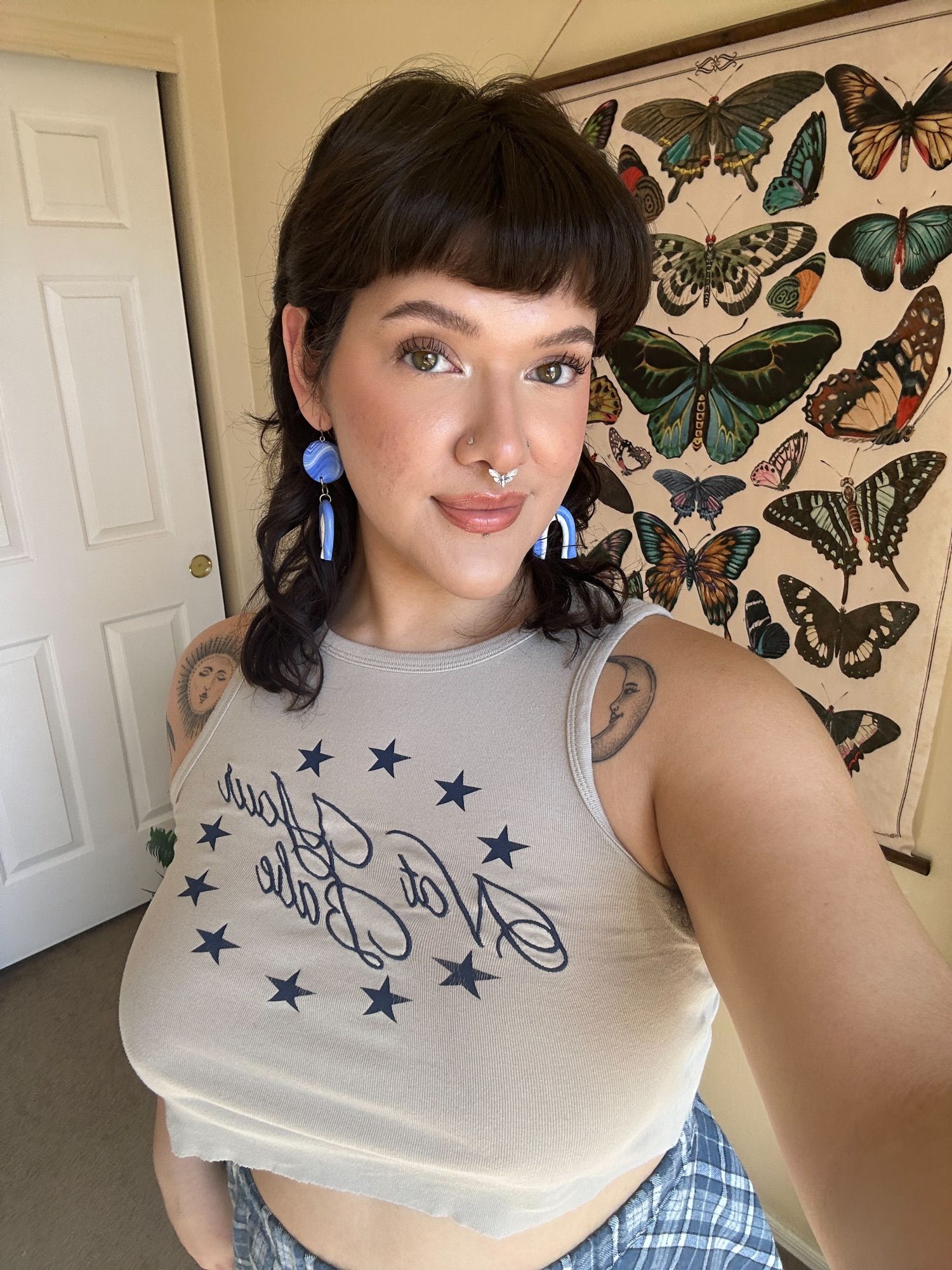 cherry taking a selfie while wearing blue & white rainbow earrings, a beige tank top that says “not your babe”, and a plaid blue and white skirt.