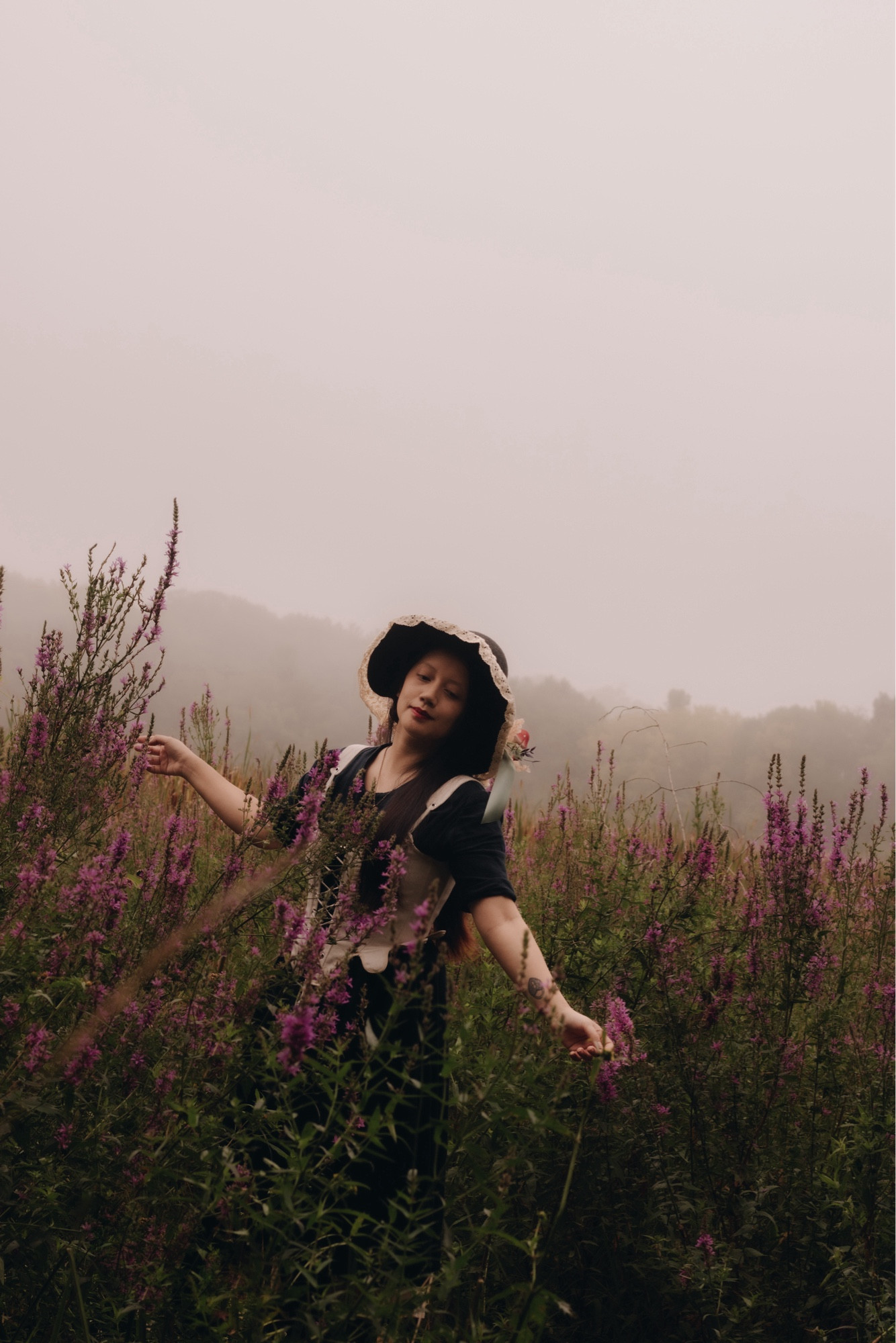 A girl, wearing a witch hat, surrounded by purple loosestrifes and fog.