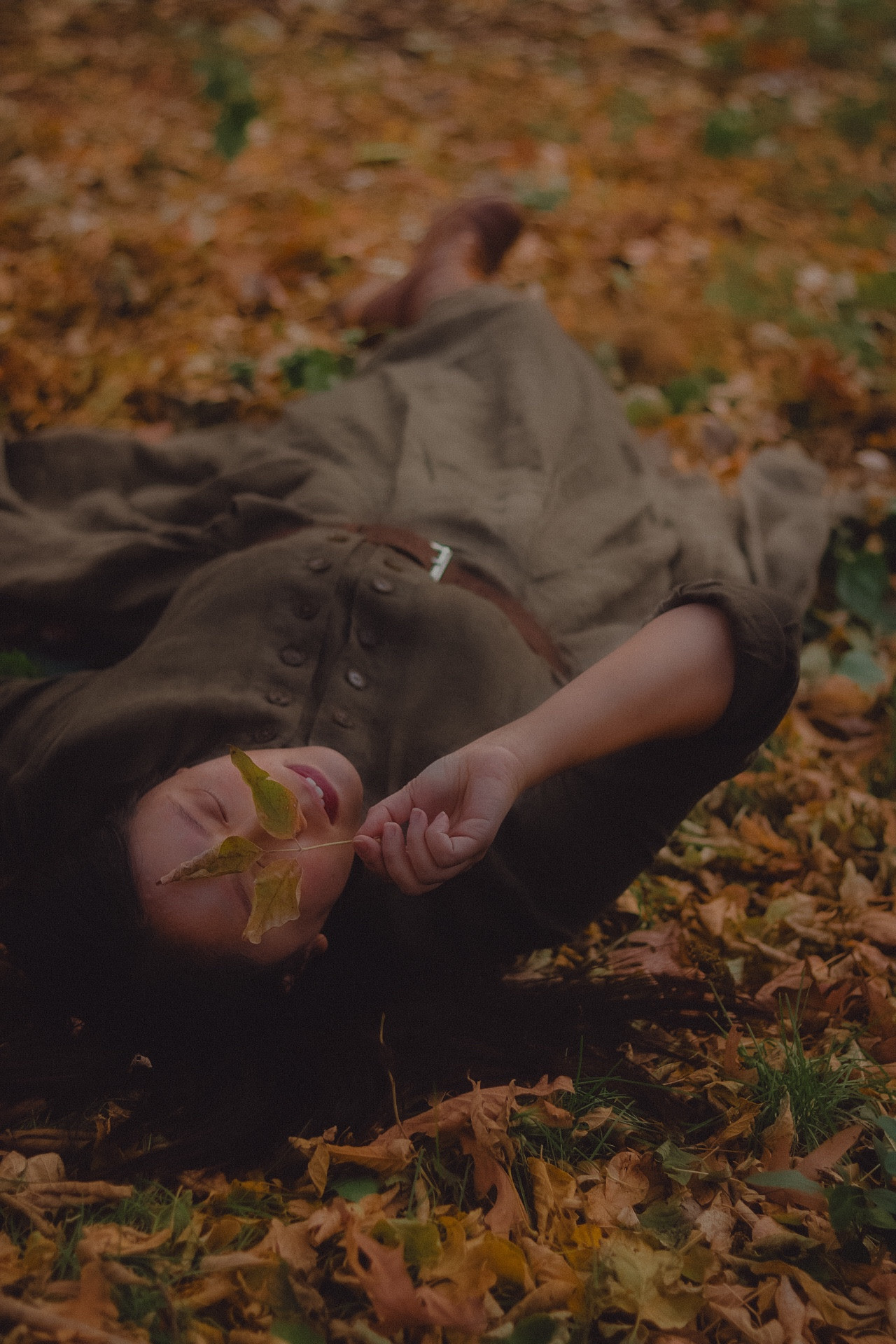 A girl laying on top of fallen leaves. She’s holding a small branch of leaves over her face.