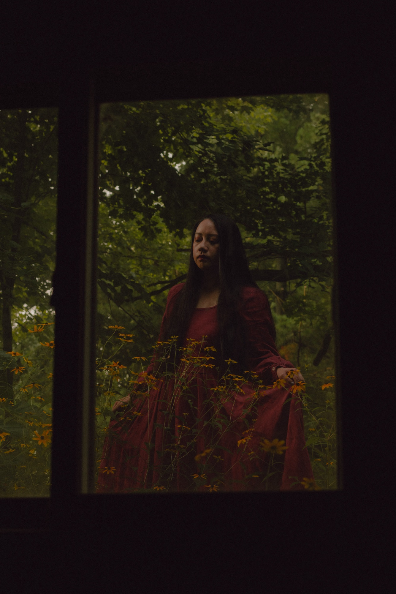 A girl behind a window. She’s wearing a red dress, standing among yellow flowers and trees.