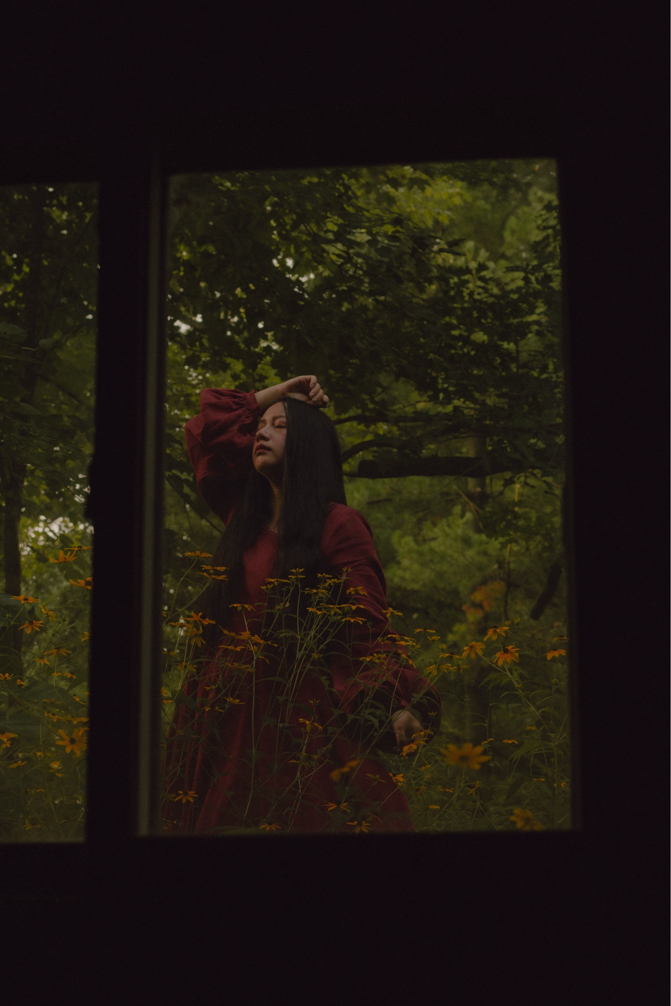 A girl behind a window. She’s wearing a red dress, standing among yellow flowers and trees.