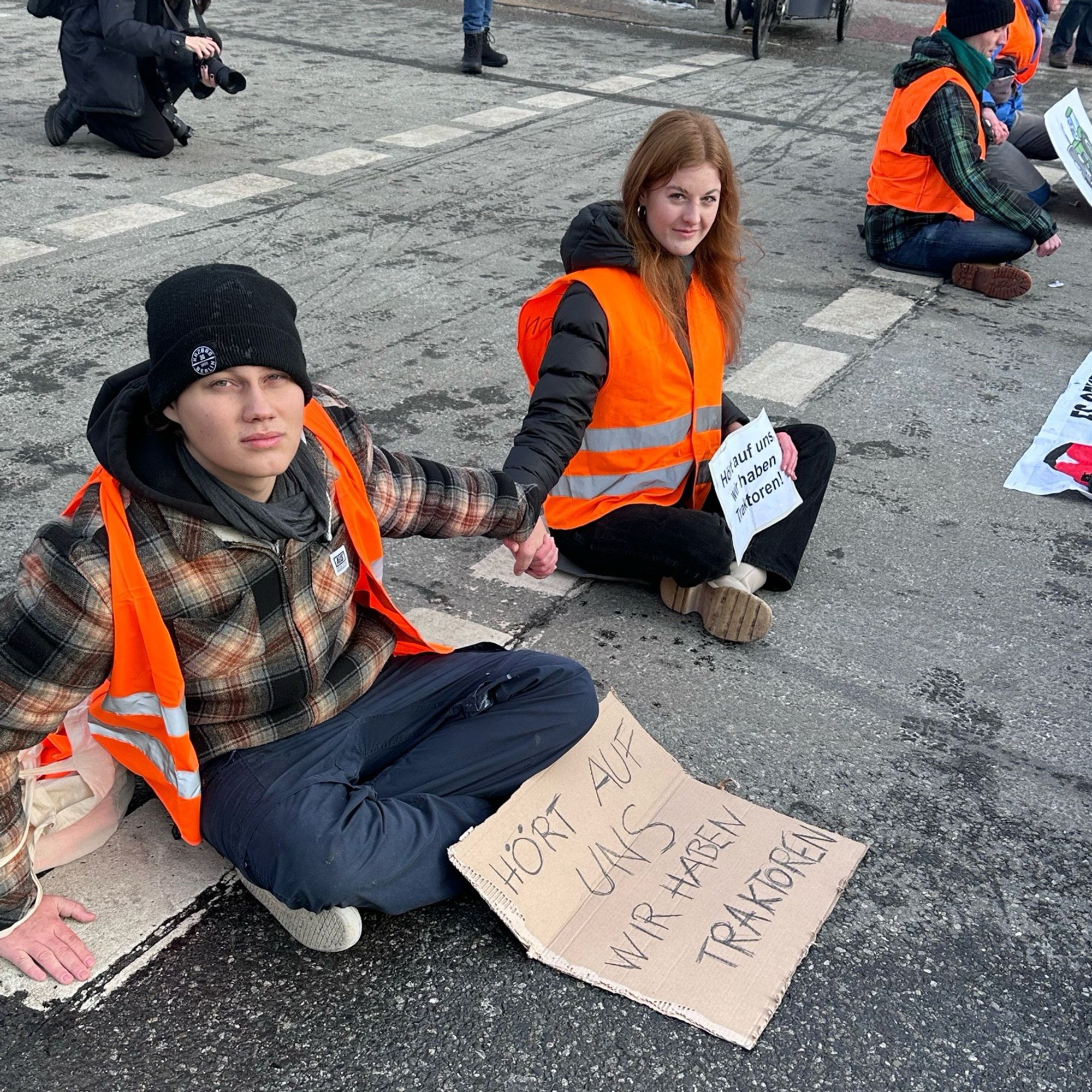 auf der Straße festgeklebte Klimaaktiviti neben Spielzeugtraktoren mit einem Schild „Hört uns zu, wir haben Traktoren“