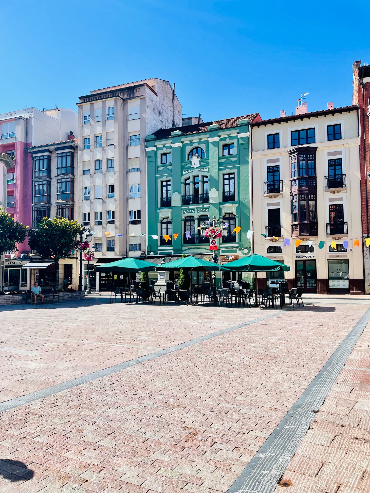 Plaza surrounded by multicoloured buildings. A cafe with table and chairs and green parasols is at the end.