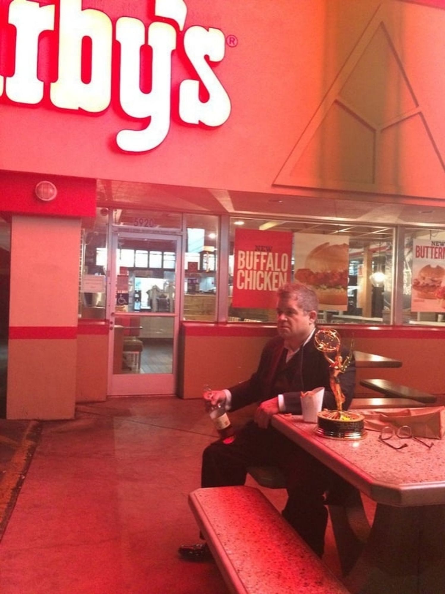 Patton Oswalt with his Emmy beside him and a bottle of liquor in his hand, staring off into the middle distance at an Arby's.