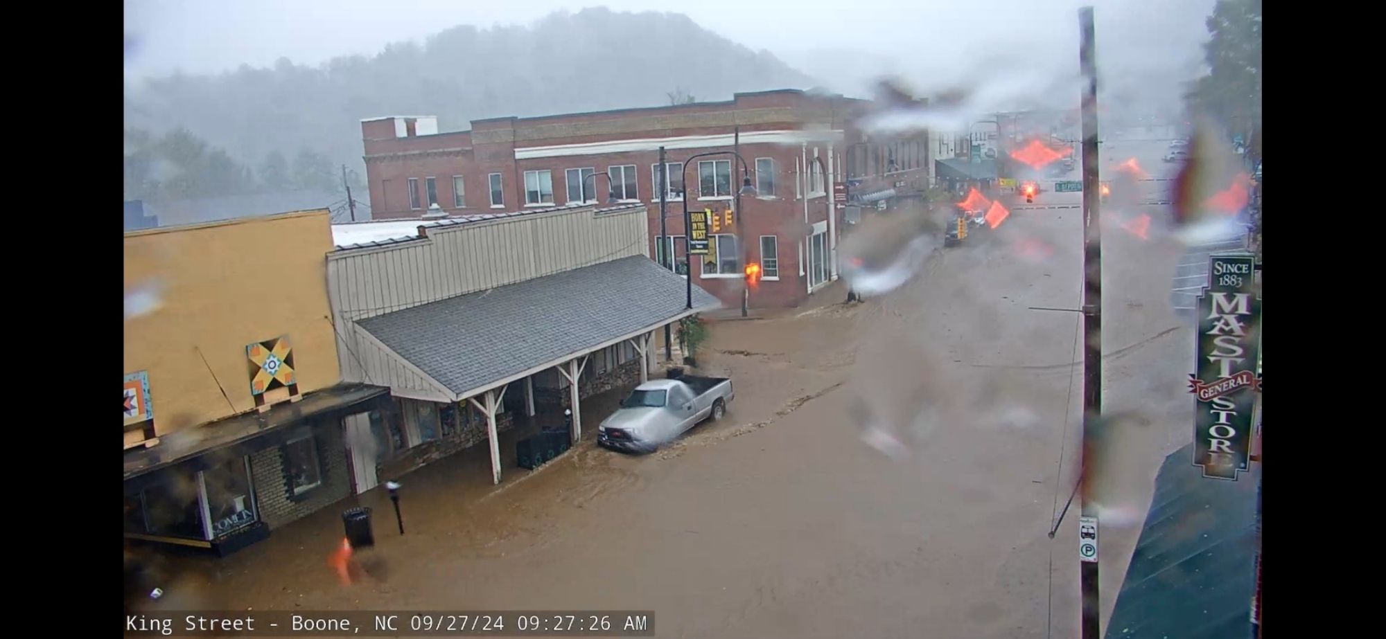 Photo of flooded downtown Boone, NC. Dirty water is covering the roadway and rising over the curb to cover the sidewalk. A pickup truck is parked and shows water rising up to near the bumper.