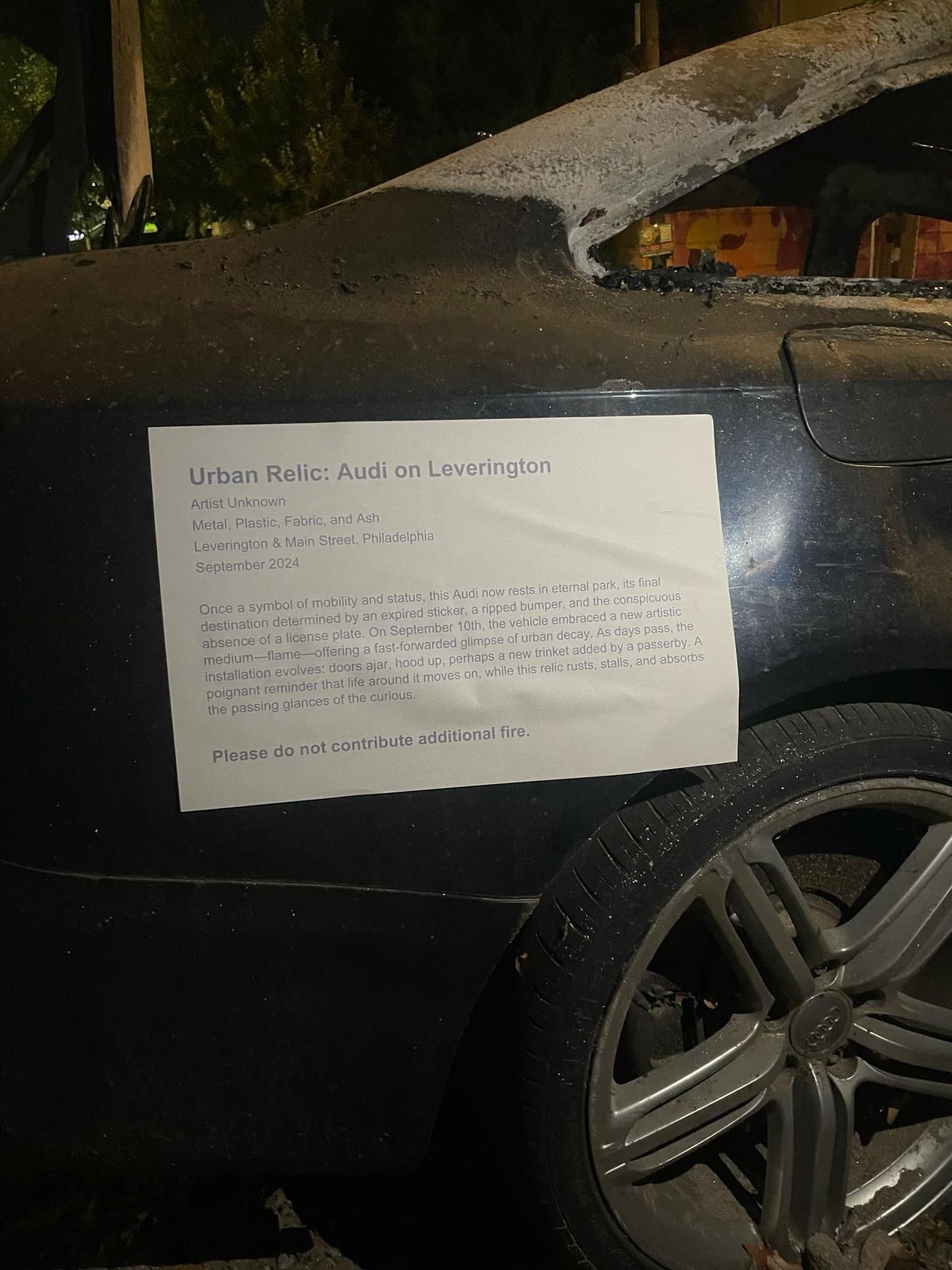 The charred remains of a burnt out car. A sign is affixed to it, which reads: 

Urban Relic: Audi on Leverington

Artist Unknown

Metal, Plastic, Fabric, and Ash

Leverington & Main Street, Philadelphia

September 2024

Once a symbol of mobility and status, this Audi now rests in etemal park, its final destination determined by an expired sticker, a ripped bumper, and the conspicuous absence of a license plate. On September 10th, the vehicle embraced a new artistic medium - flame - offering a fast-forwarded glimpse of urban decay. As days pass, the installation evolves: doors ajar, hood up, perhaps a new trinket added by a passerby. A poignant reminder that life around it moves on, while this relic rusts, stalls, and absorbs the passing glances of the curious.

Please do not contribute additional fire.