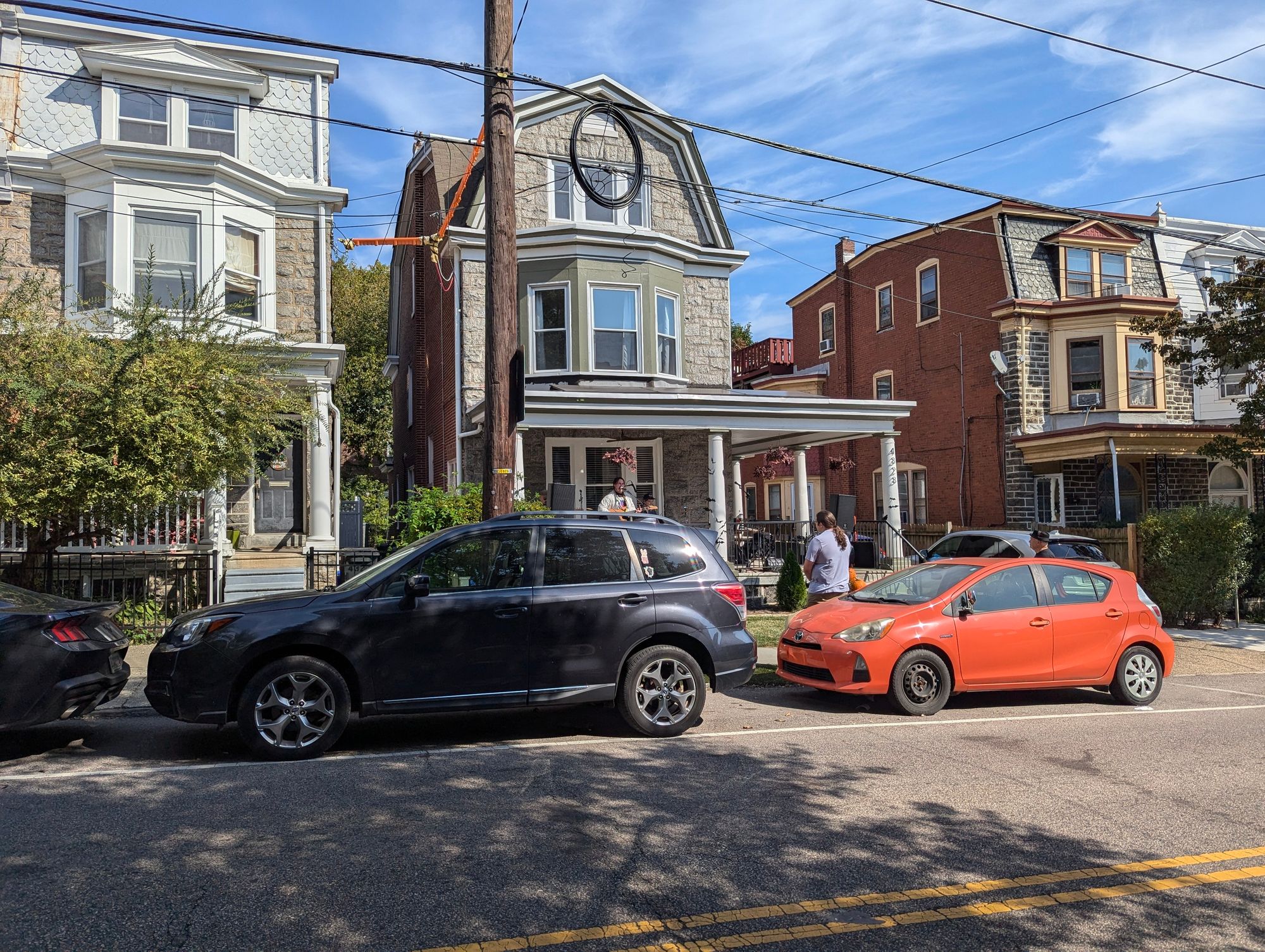 The image captures a vibrant street scene under a clear blue sky. Dominating the foreground is a black car, parked neatly by the side of the road. Its sleek form contrasts with the red car on its right, which sports a white roof and a silver spoiler, adding a touch of sportiness to the scene.

In the background, three more cars are visible. Two of them are as black as night, while one stands out in a striking shade of orange. They are parked in front of a brick building, which boasts a porch adorned with columns, lending an air of elegance to the otherwise ordinary structure.

The street itself is lined with trees, their lush green leaves forming a natural canopy overhead. Their presence adds a touch of nature to this urban setting. The precise location of these elements creates a harmonious balance in the image, making it not just a simple snapshot of cars on a street, but a scene that tells a story of everyday life and city living.