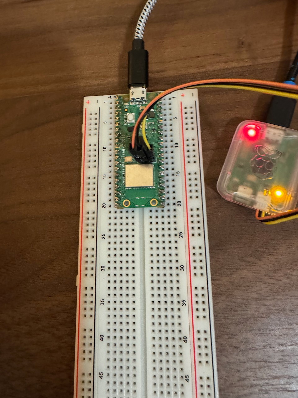 Raspberry Pi Pico W connected to a breadboard with jumper wires attached, wired to a Raspberry Pi Debug Probe emitting red and yellow LEDs. The setup is displayed on a wooden desk, showcasing an active debugging or prototyping configuration.