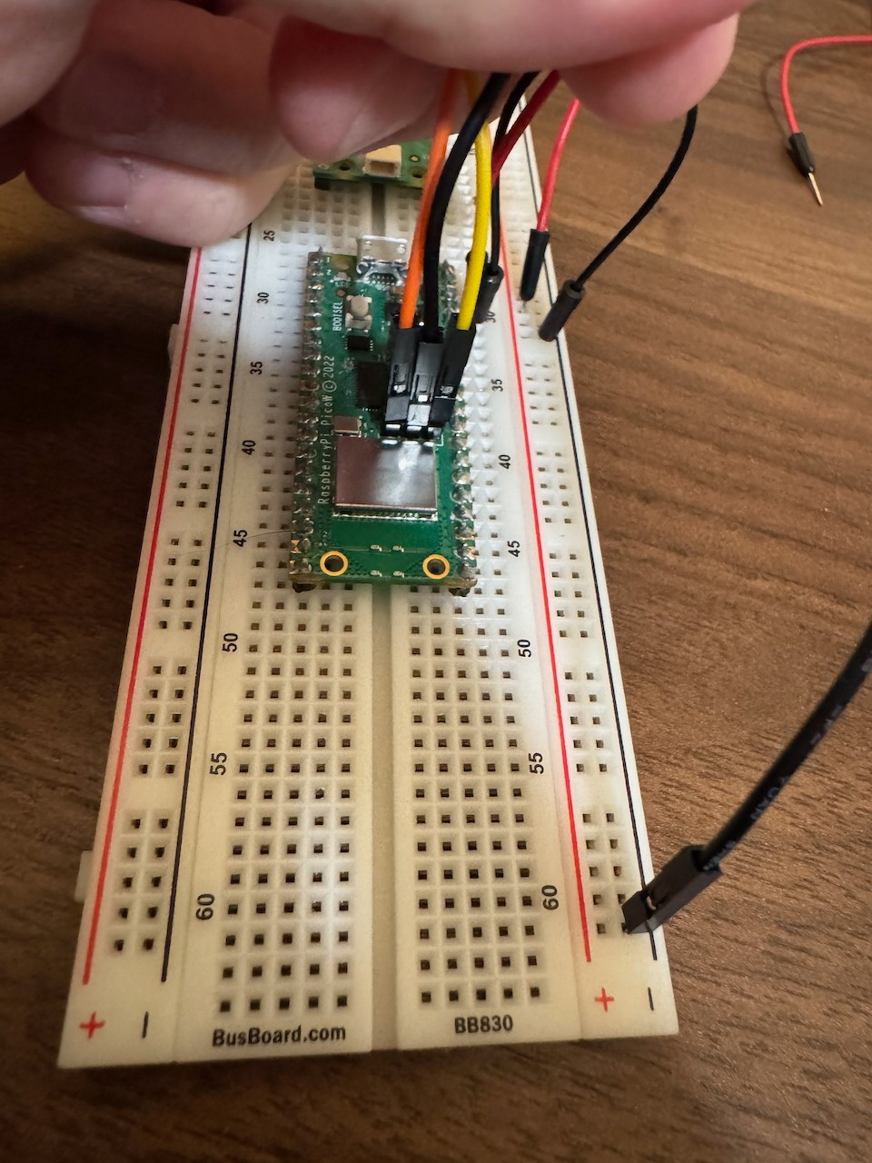 View of a Raspberry Pi Pico W mounted on a breadboard, with jumper wires in red, orange, yellow, and black being connected by hand. The close-up emphasizes the wiring process in an electronics prototyping setup, with additional wires scattered on the desk.