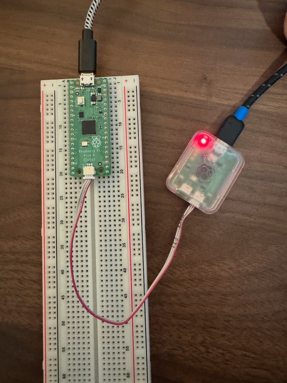 Close-up of a Raspberry Pi Pico connected to a breadboard, wired to a Raspberry Pi Debug Probe with a glowing red LED, indicating active status. The setup is placed on a wooden desk, showing a simple hardware debugging configuration.