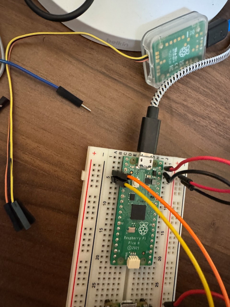 Close-up of a Raspberry Pi Pico H mounted on a breadboard, wired to a Raspberry Pi Debug Probe visible in its case, with connected jumper wires in red, orange, and yellow. The setup showcases active wiring for debugging or interfacing. Other wires and components are visible in the background on the wooden desk.
