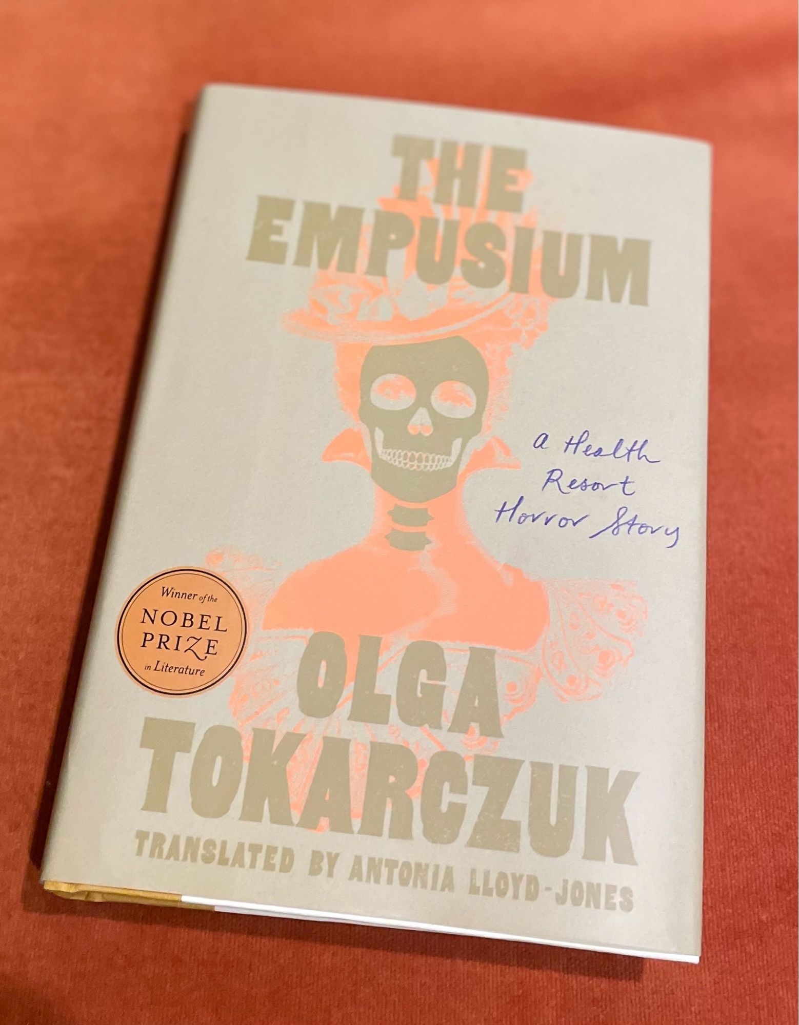 Olga Tokarczuk’s most recent book EMPOSIUM: A HEALTH RESORT HORROR STORY translated into English by Antonia Lloyd-Jones. Hardcover has a creepy greenish brown skull wearjng a pastel orange Victorian hat & high-necked dress. Orange sticker says “Winner of the NOBEL PRIZE in Literature.”