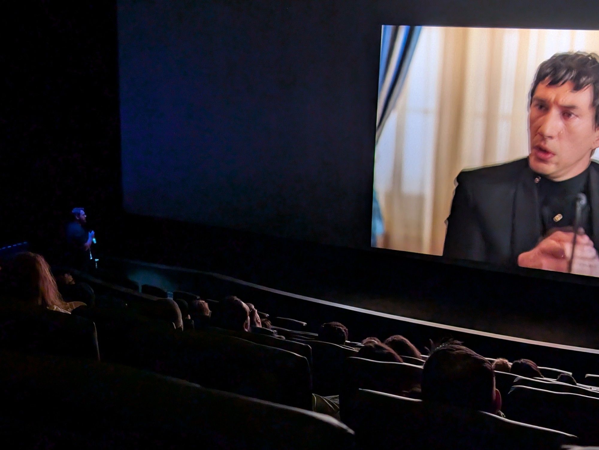 A man with a microphone addresses an IMAX screen with Adam Driver on it at a showing of Megalopolis