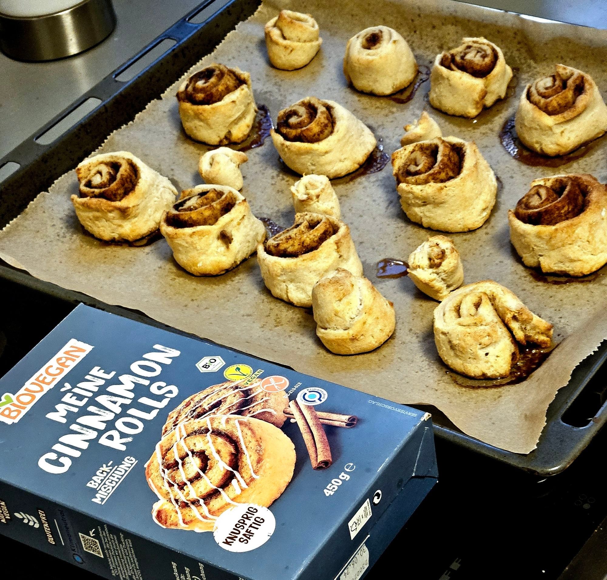 Ein Backblech mit vielen etwas unförmigen Zimtschnecken, daneben eine leere Packung "Meine Cinnamon Rolls" Backmischung von biovegan.