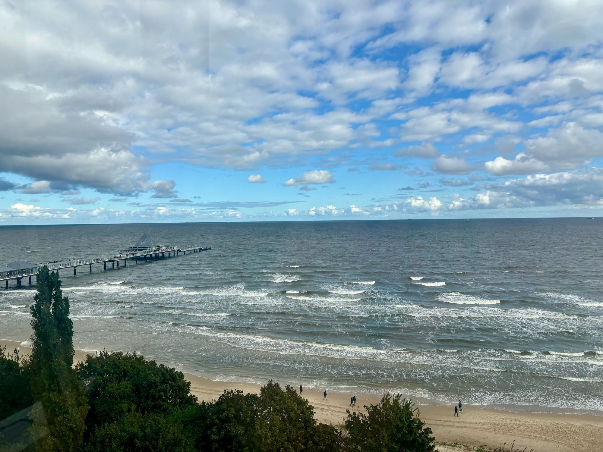 Blick aus der Höhe auf die Ostsee. Am Strand gehen Menschen spazieren, sie sehen sehr klein aus. Links ragt die Seebrücke Heringsdorf ins Bild und Meer. Am Wellengang ist zu erkennen, dass es windig ist. Der Himmel ist bewölkt, das hellblau scheint dennoch durch.