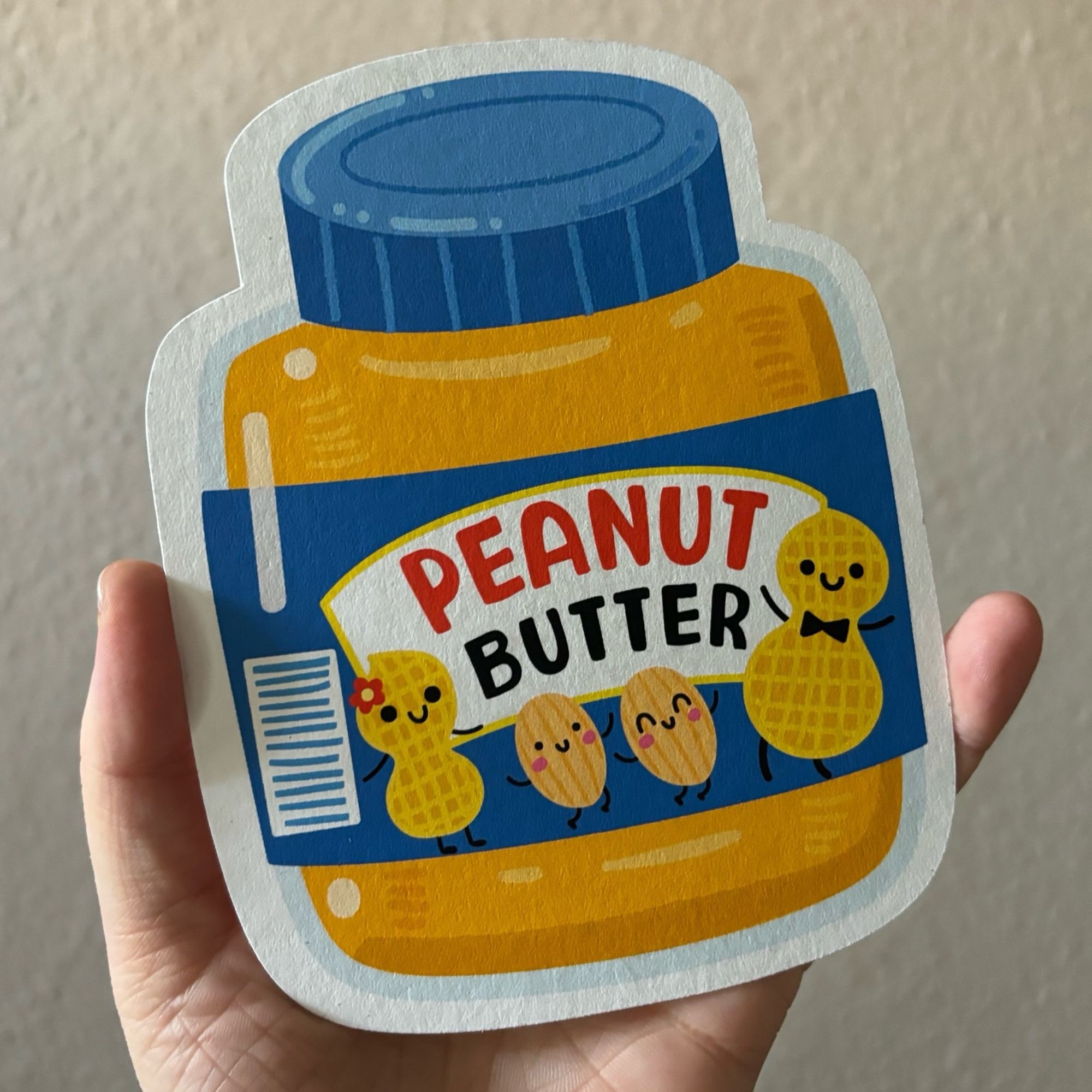 Ich halte eine Postkarte in meiner Hand, die die Form eines Glases Erdnussbutter hat. Sie ist liebevoll illustriert. Auf dem Label ist eine fröhliche Erdnuss-Familie abgebildet.