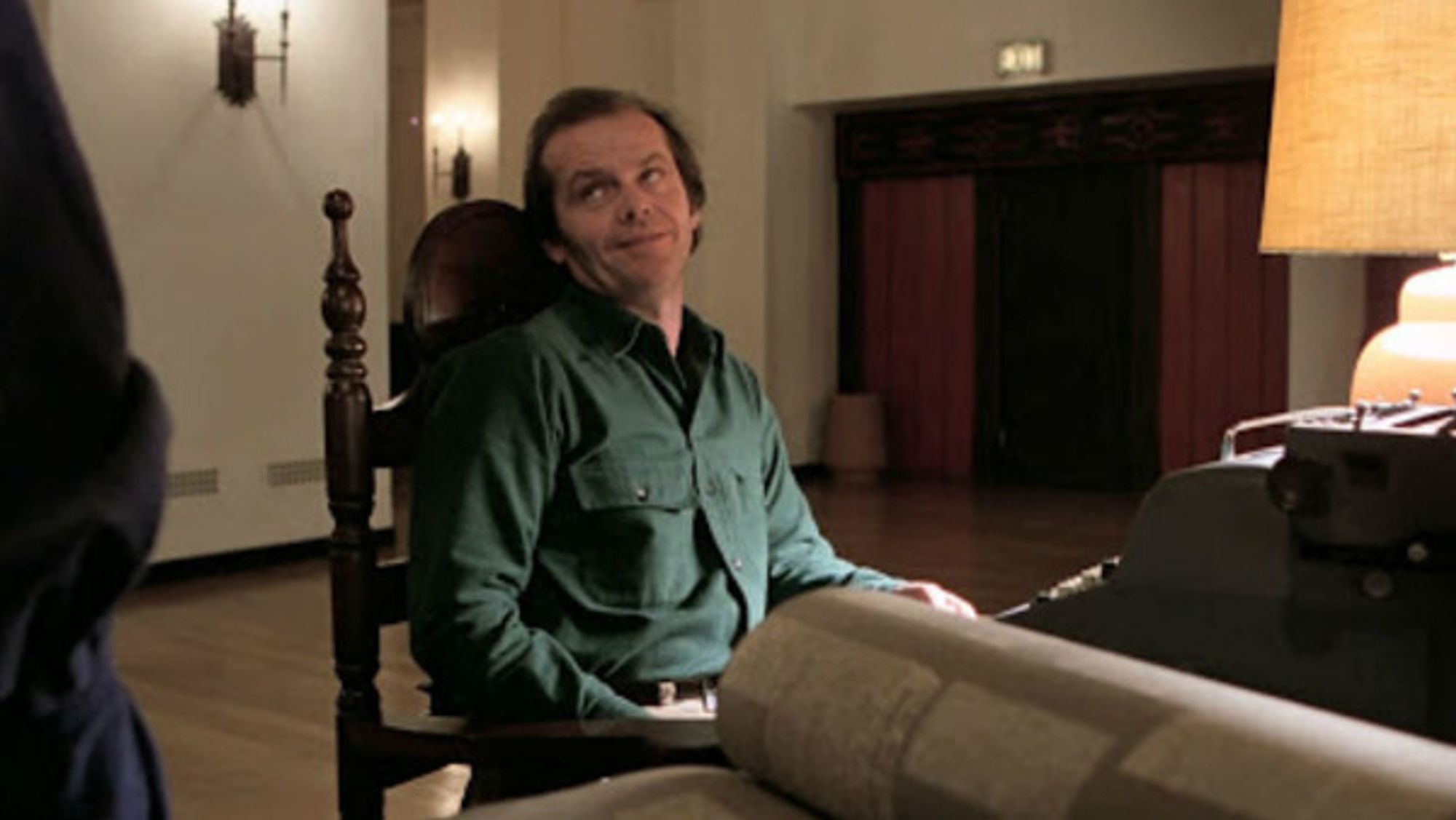 Jack Nicholson as Jack Torrance in The Shining - a middle-aged white guy sitting on an old chair at a desk in an otherwise empty hotel lobby; a typewriter is on the desk in front of him