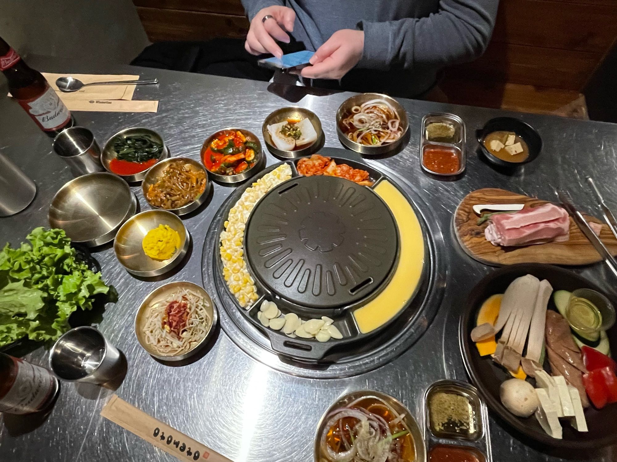 Looking down at a table top grill surrounded by cheese corn, kimchi, and egg, plus five side dishes, lettuce, pork belly, and vegetables. Seated at the table is a man holding his smartphone. There is a beer on the table next to some utensils. 