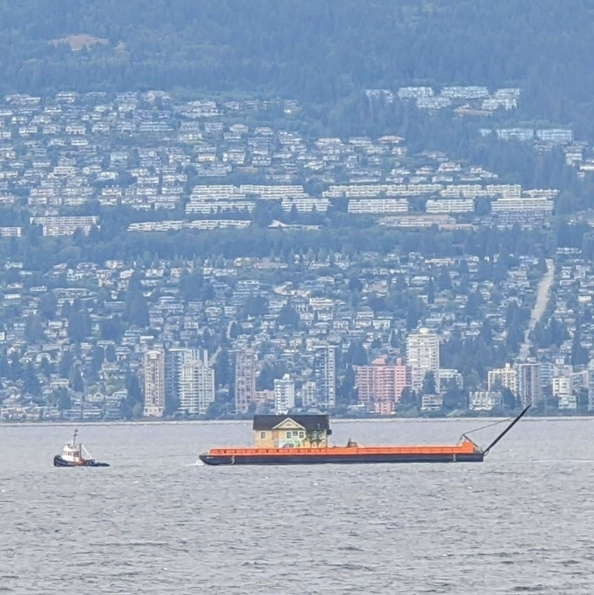 Against the backdrop of West Vancouver, the old Henry Hudson schoolhouse is pulled across English Bay on a big barge by a tug.