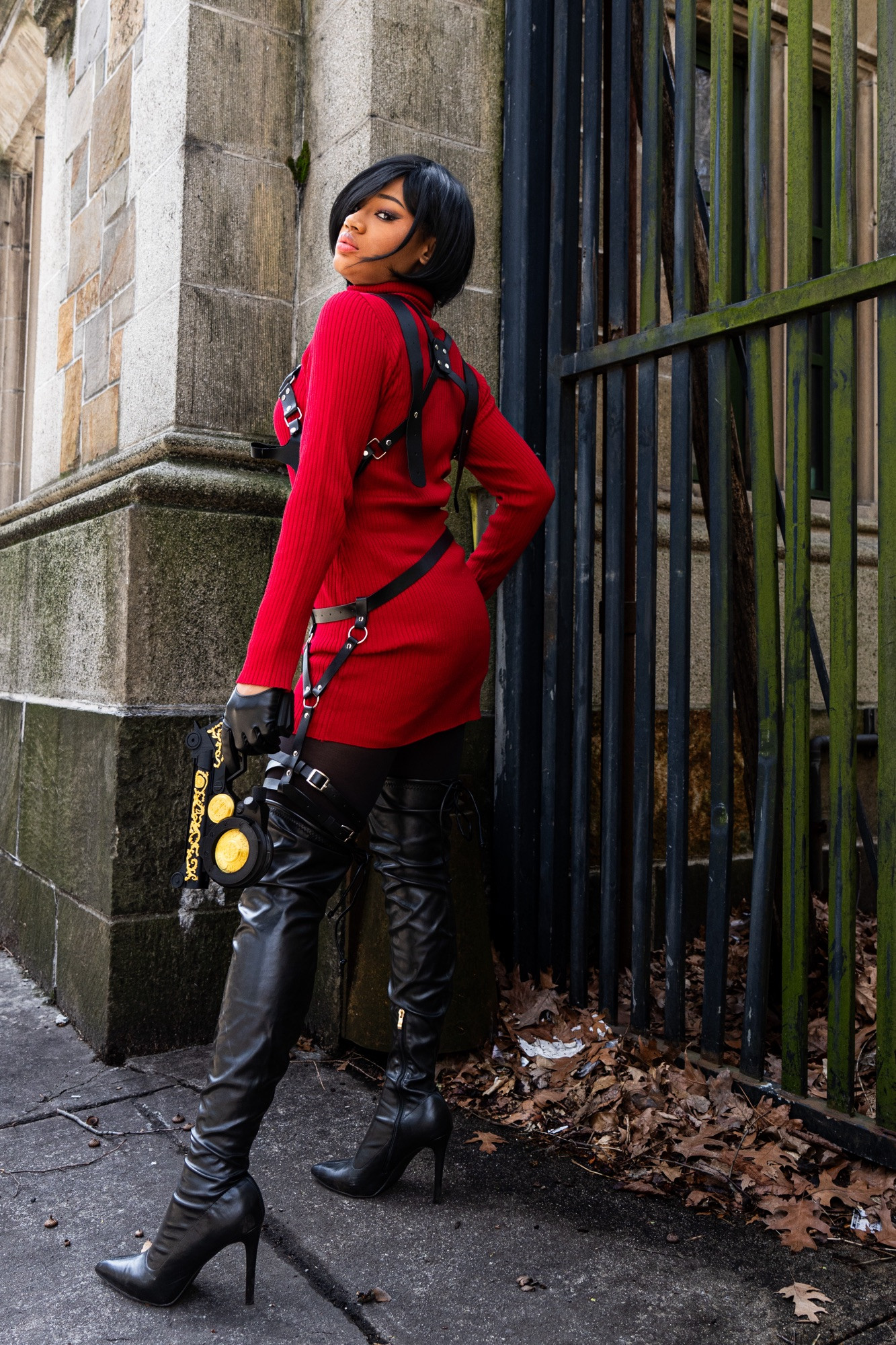 A black cosplayer is dressed as Ada Wong from resident evil 4 wearing a red sweater dress with thigh high leather heeled boots. They hold her black and gold hookshot with leather harnesses. 