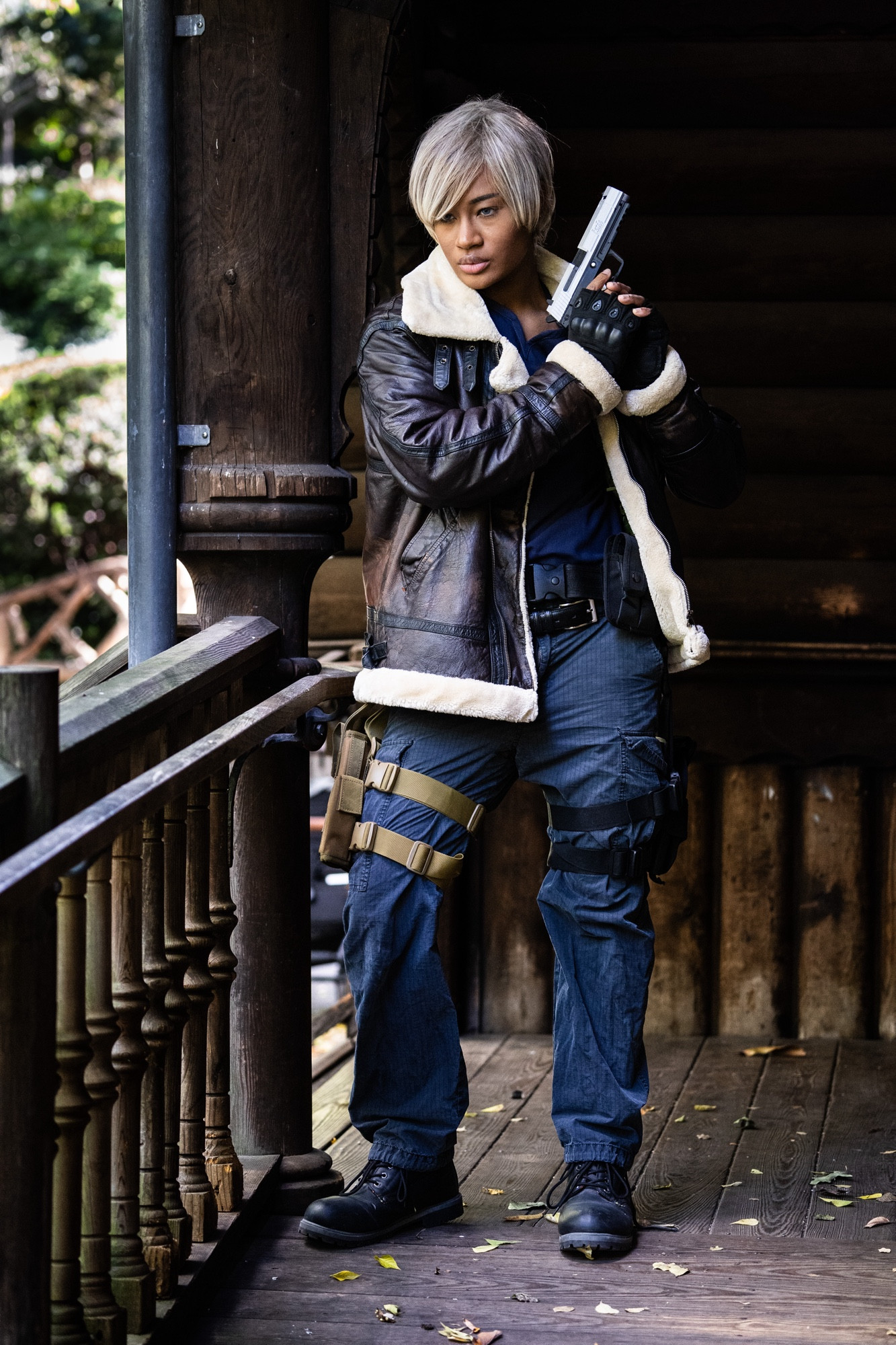 A Black person is dressed as Leon Kennedy from the Resident Evil 4 Remake. They wear a brown shearling jacket with a blue shirt under, gray pants and two gun holsters on their leg with combat boots. They have a blond wig on with blue eyes and stand on a run down cabin’s front porch. They hold a prop up to their head looking to their left